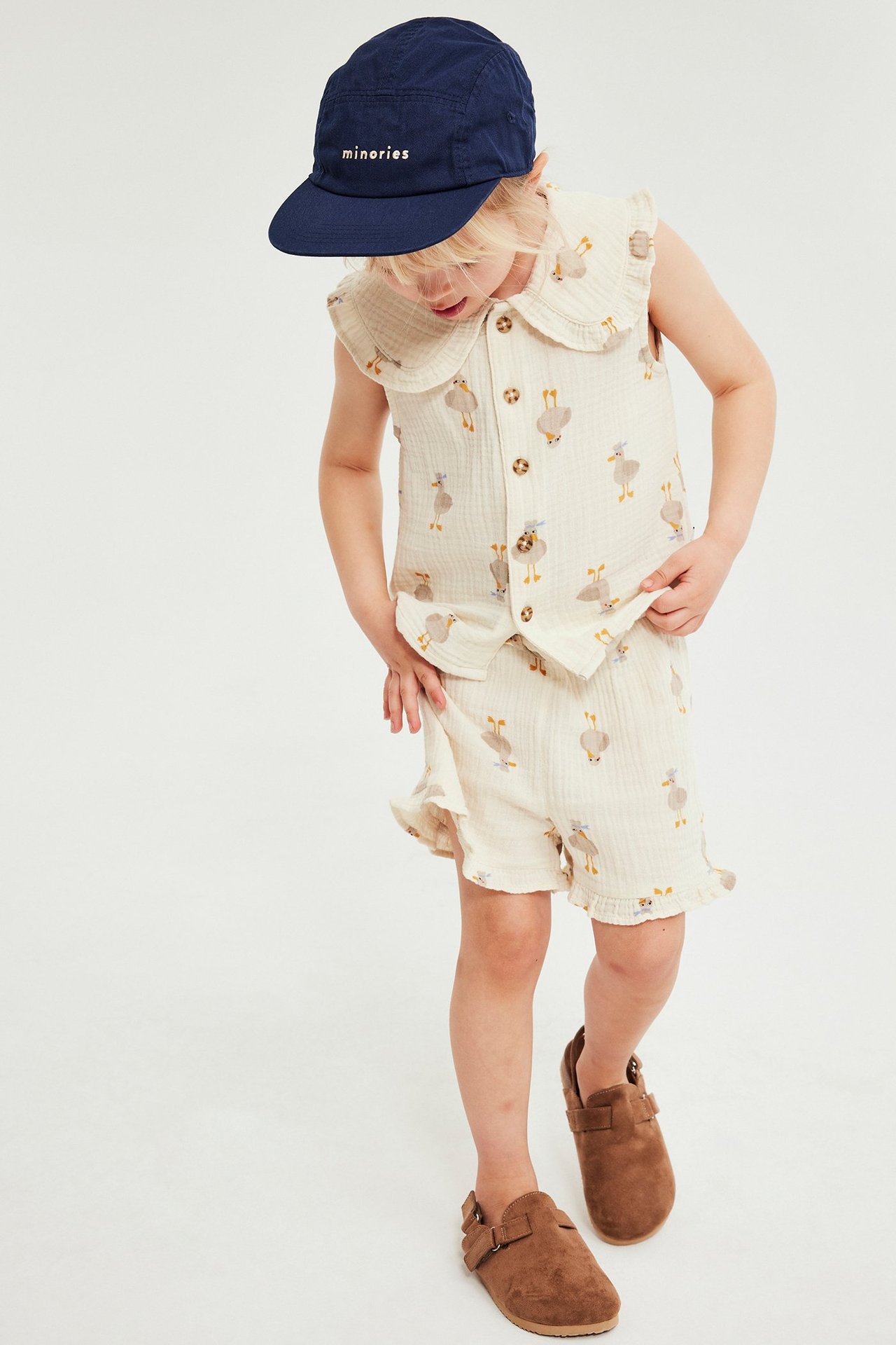 Framifrån: Barn i beige mönstrat bomullslinne och shorts-set, marinblå keps och bruna skor.