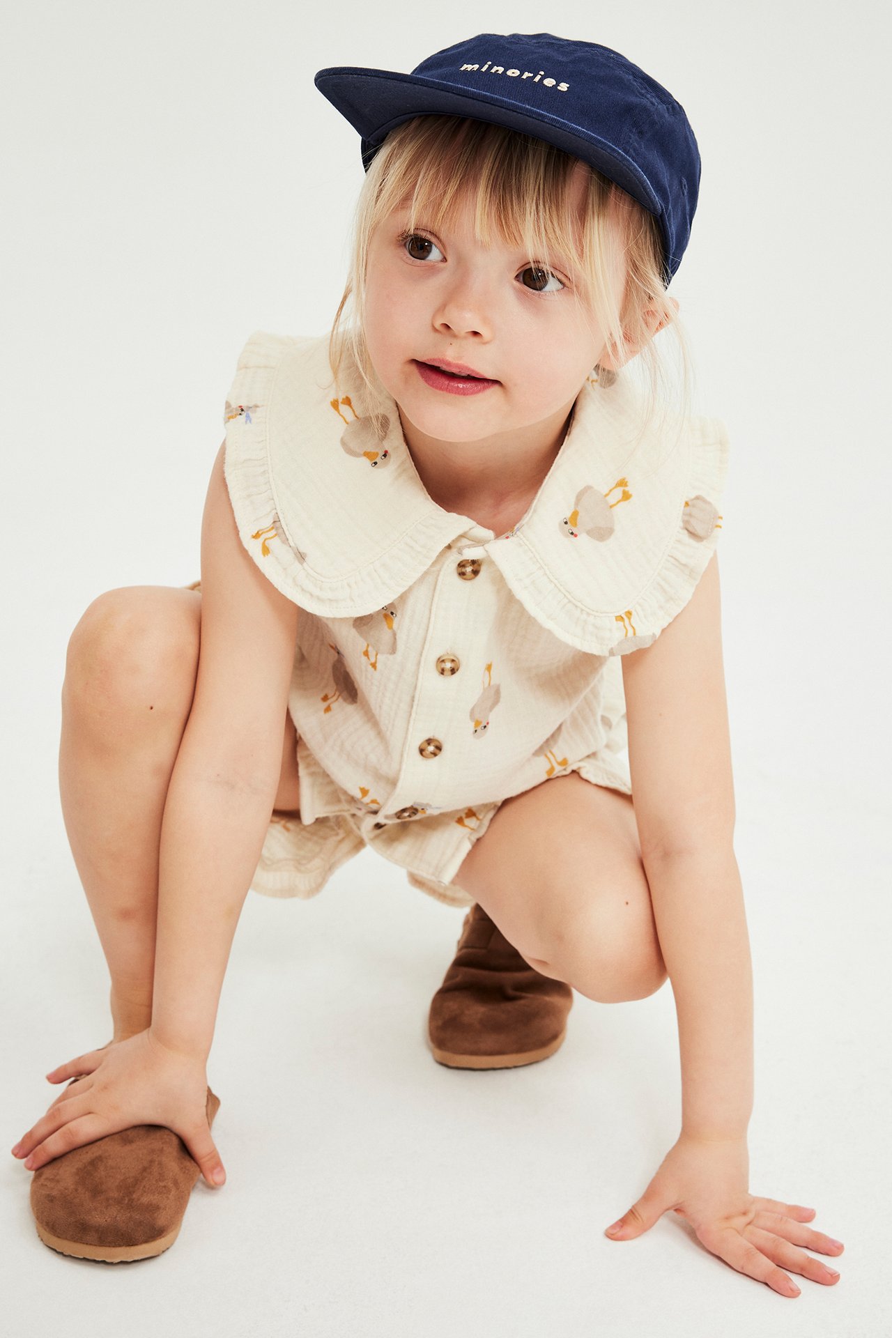 Frontvy: Ett barn i beigefärgat mönstrat vävt bomullsset med volangkrage topp och shorts, plus mörkblå keps.