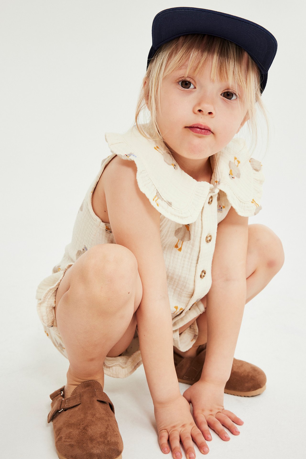 Kid bär ett mönstrat beige bomulls-set med linne, shorts, volangkrage, marinblå keps och bruna tofflor.