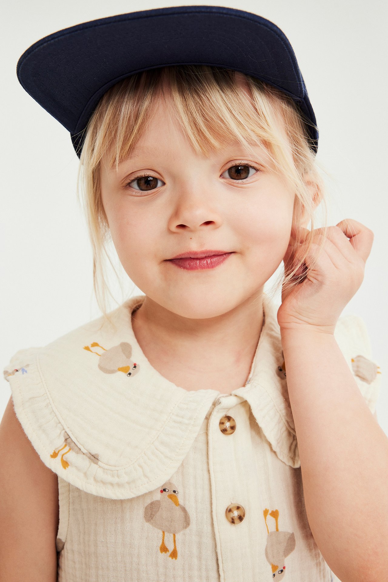 Närbild: Kid flicka i beige bomullslinne med fågelmönster, knappar fram och marinblå keps.