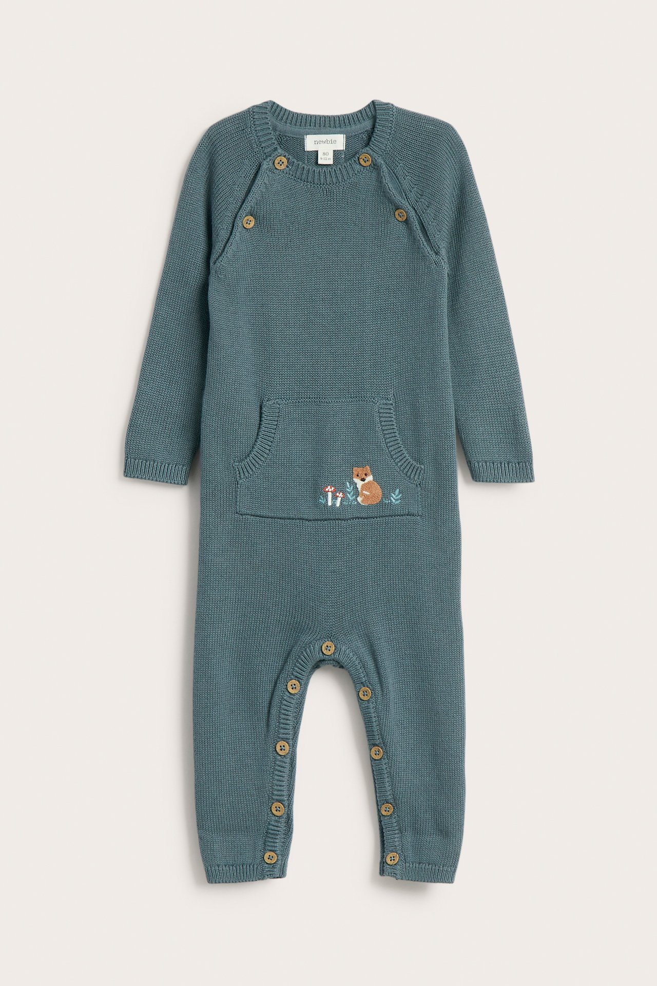 Front av en strikket babyoverall i petroleumsblå med knapper og brodert reve-lomme.