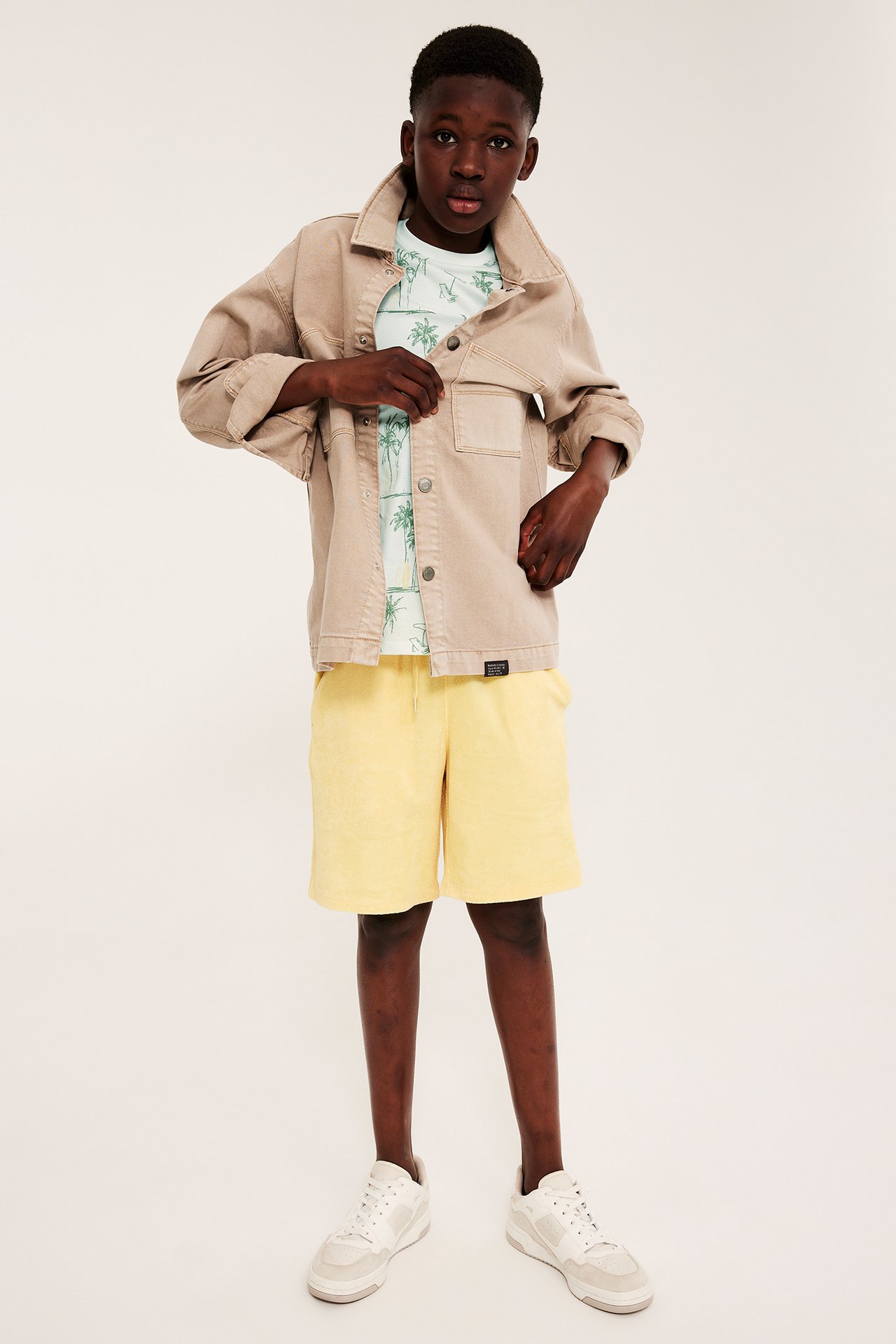 Frontvy av ett barn i offwhite t-shirt med palmmönster, beige jacka, gula shorts och vita sneakers.