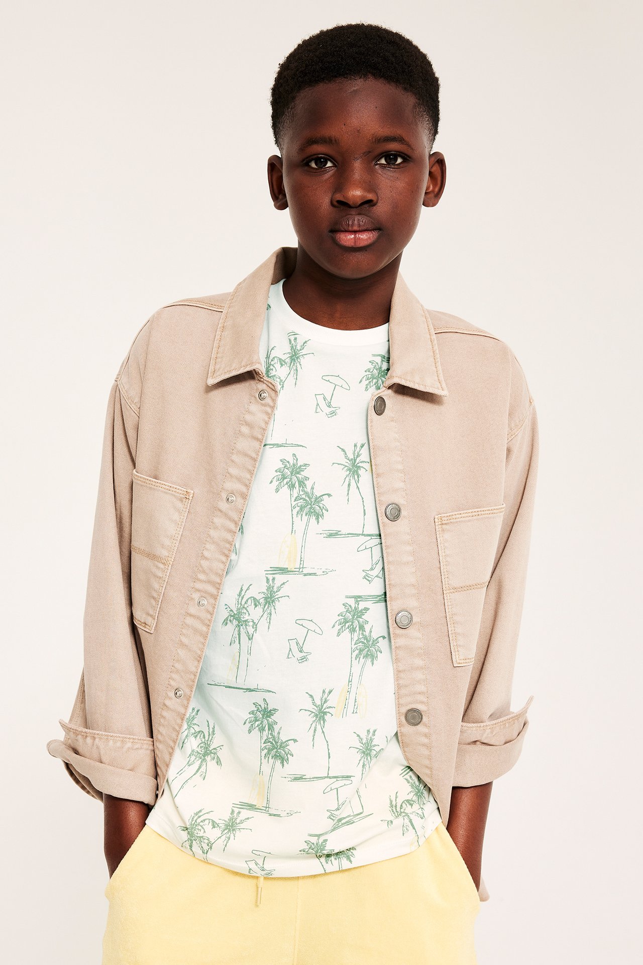 Frontbild på ett barn i offwhite t-shirt med palmtryck, beige jacka och gula shorts.