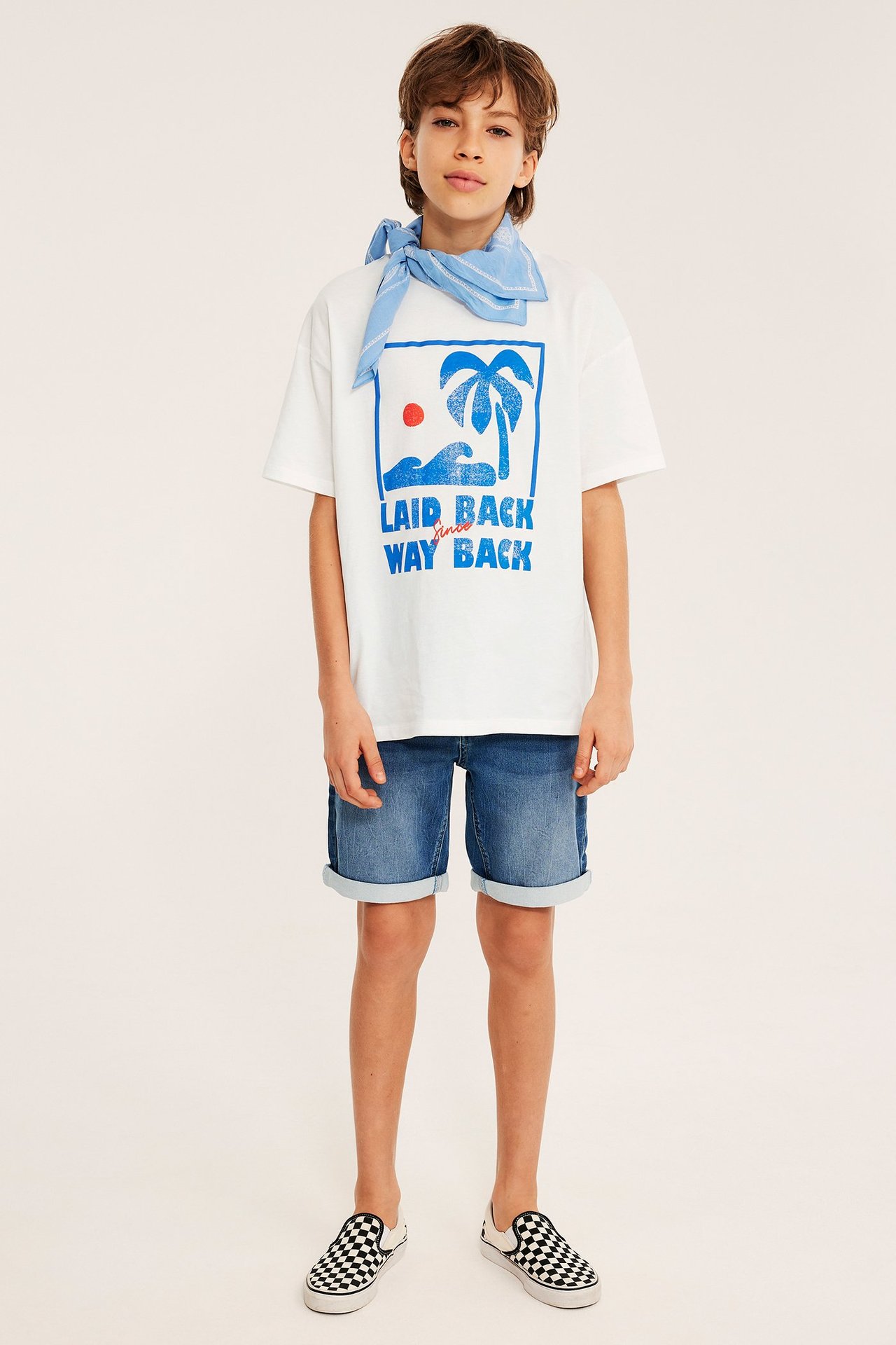 Barn bruker off-white oversized t-skjorte med blått trykk, blå denimshorts, lyseblå bandana, rutete sko.