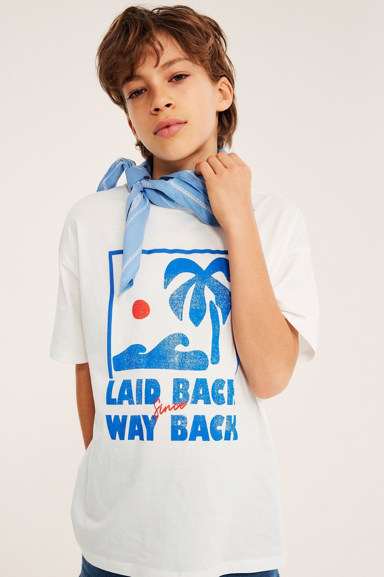 Etukuva lapsesta yllään luonnonvalkoinen oversized puuvilla t-paita sinisellä rantaprintillä ja vaaleansininen huivi.