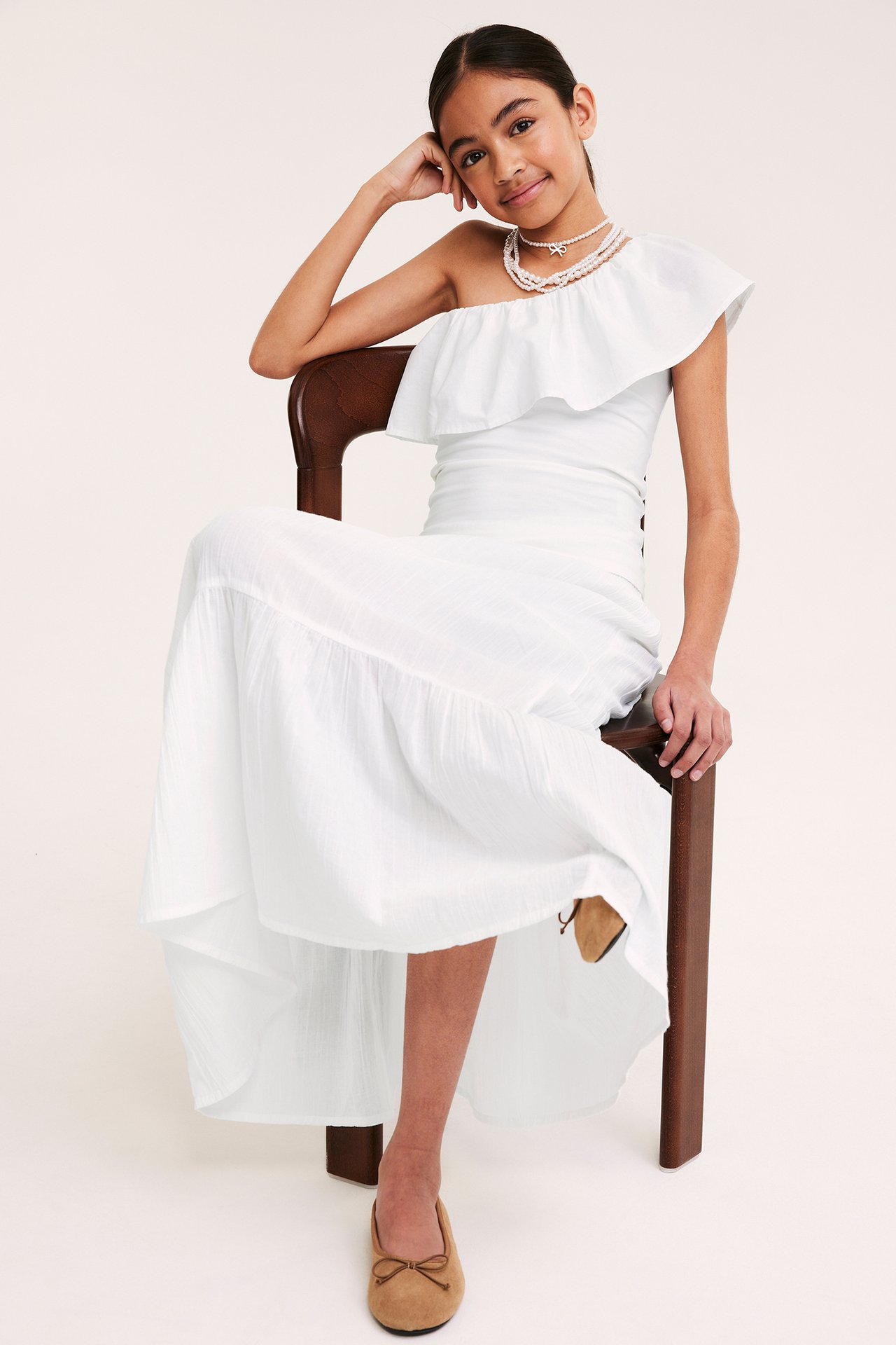 Framsida på barn i vit volangkantad bomullsklänning med en axel, pärlhalsband och beiga ballerinaskor.