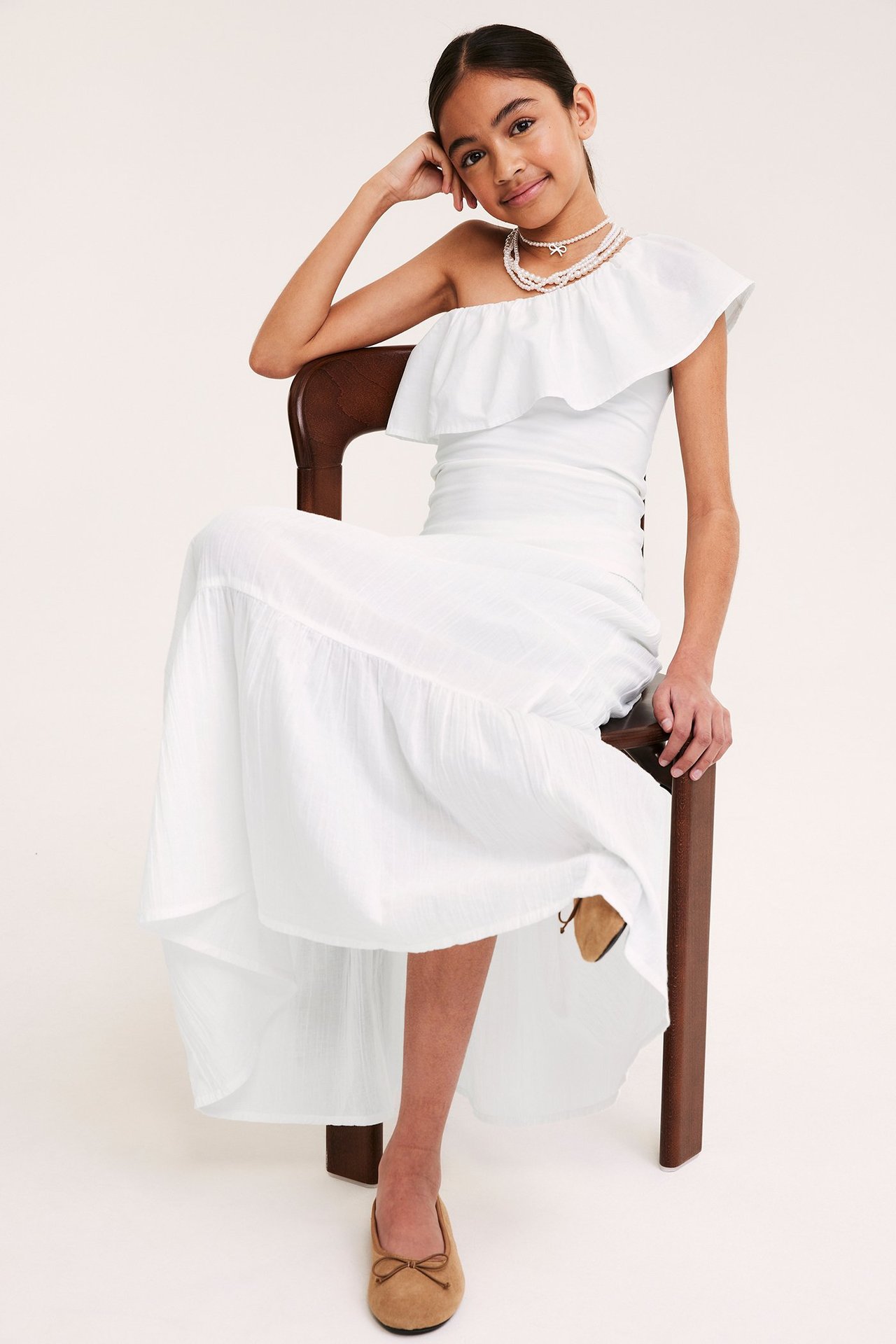 Framsida på barn i vit volangkantad bomullsklänning med en axel, pärlhalsband och beiga ballerinaskor.