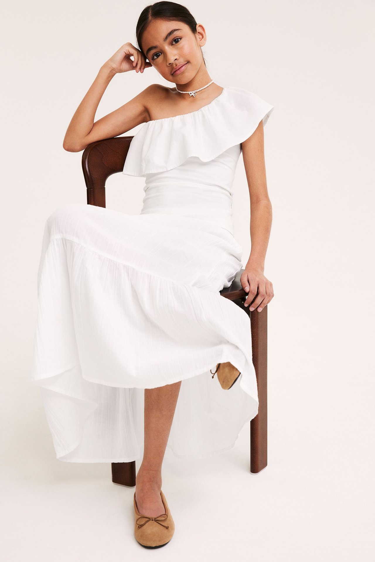 Framifrån: Barn i vit volangklänning med en axel, halsband och ljusbruna ballerinaskor.