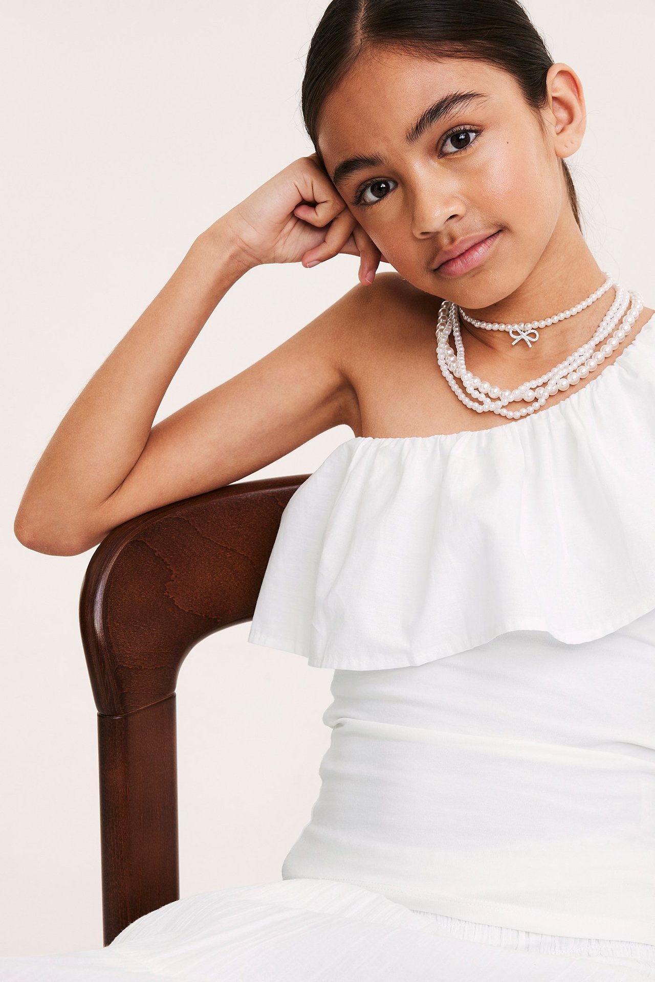 Frontvy: En flicka bär en vit bomullstopp med en axel och volang samt ett pärlhalsband.