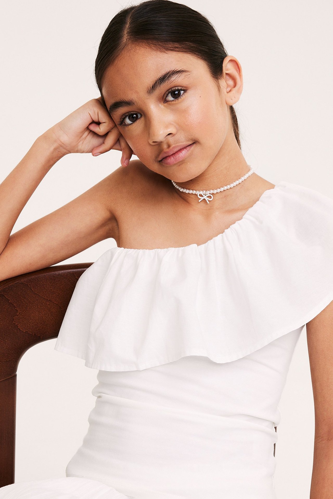 Framsida på ett barn i vit enaxlad volangtopp av bomull och pärlhalsband med rosett.