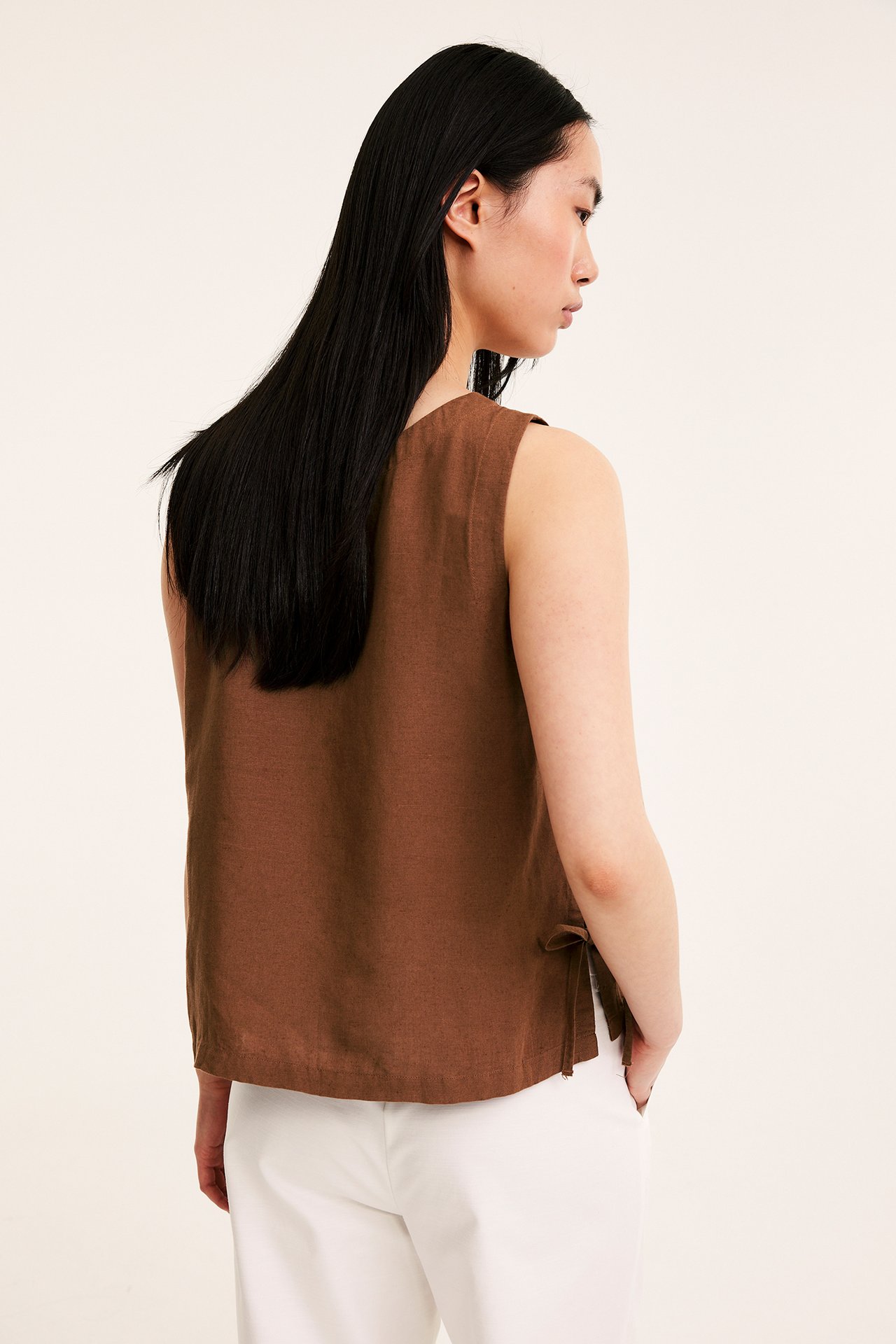 Widok kobiety z boku-tyłu: brązowy top z mieszanki lnu z wiązaniami po bokach i białymi spodniami.