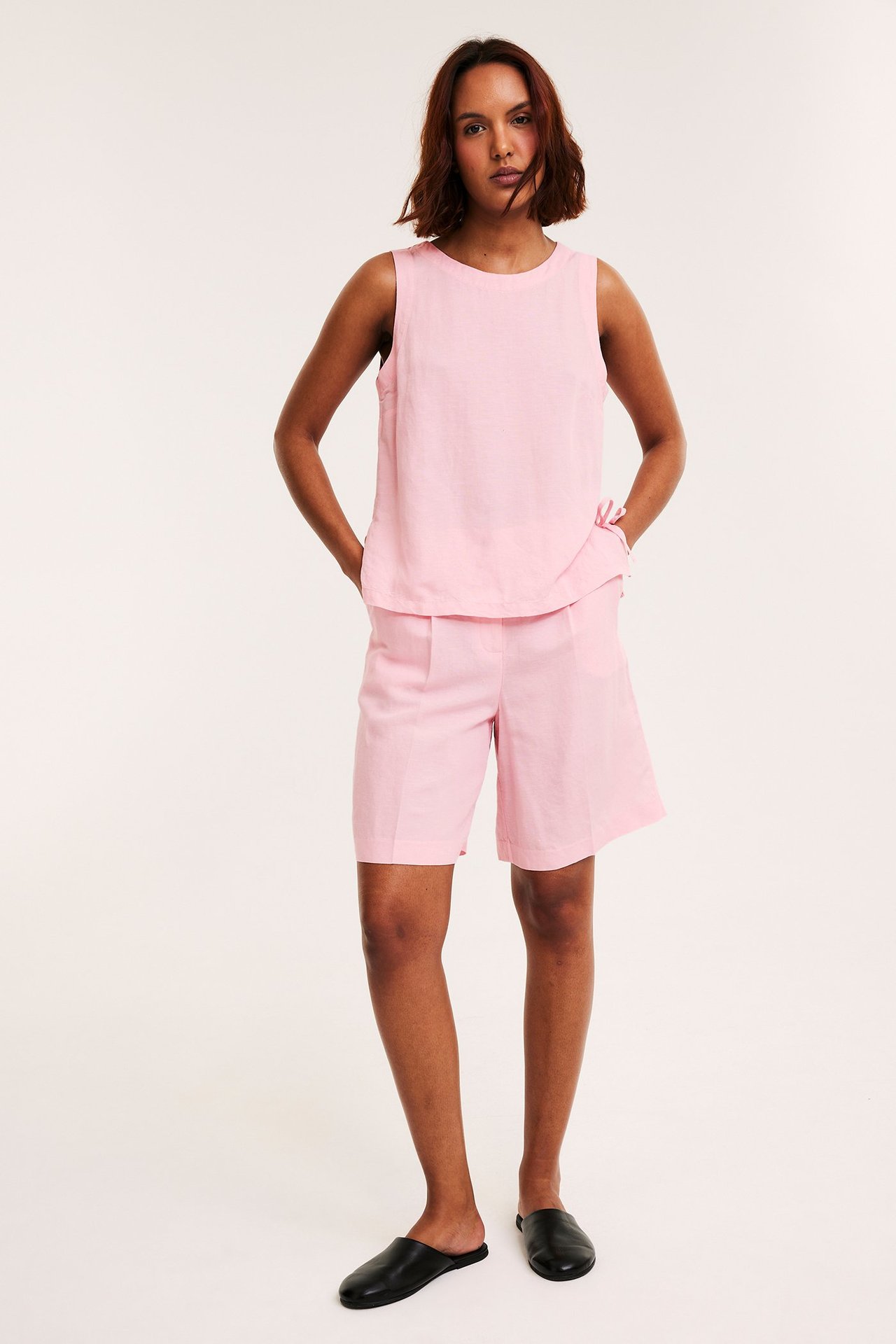 Forfra: kvinne i lys rosa linblandingstopp med knytebånd i siden, matchende shorts og svarte slip-ons.