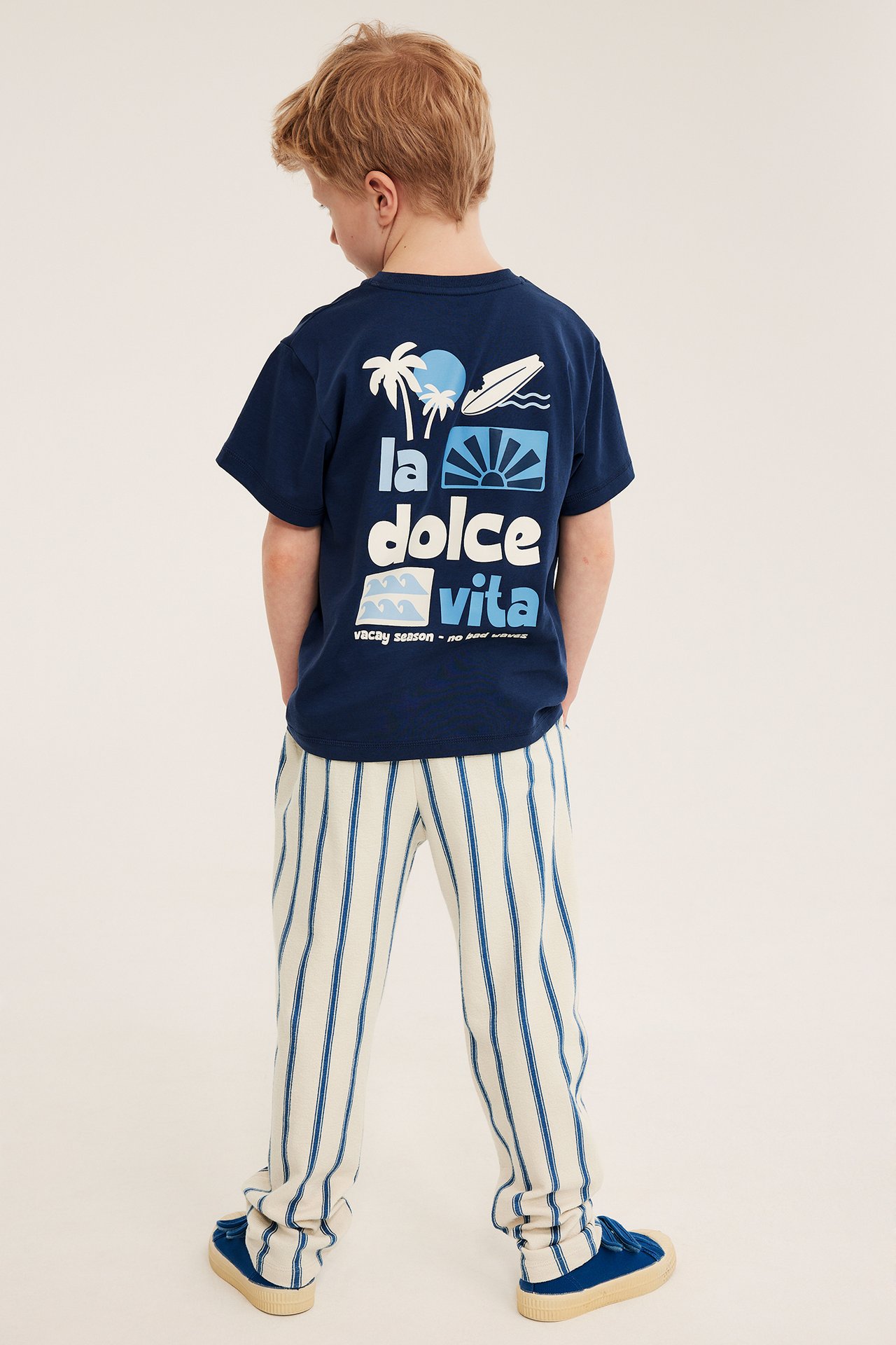 Ryggvy av ett barn i ljusbeige randiga joggingbyxor av bomull, en marinblå t-shirt och blå skor.