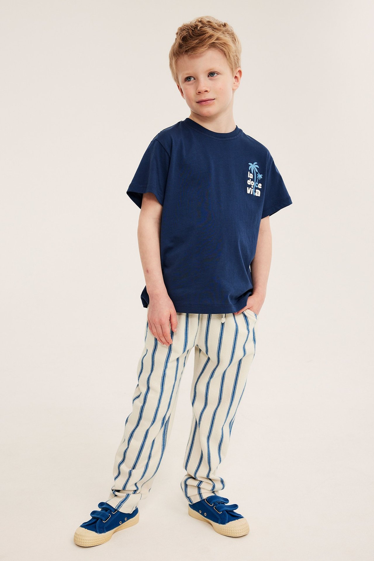 Framifrån, ett barn i beiga randiga bomullsjoggingbyxor, en marinblå t-shirt och blå tygskor.