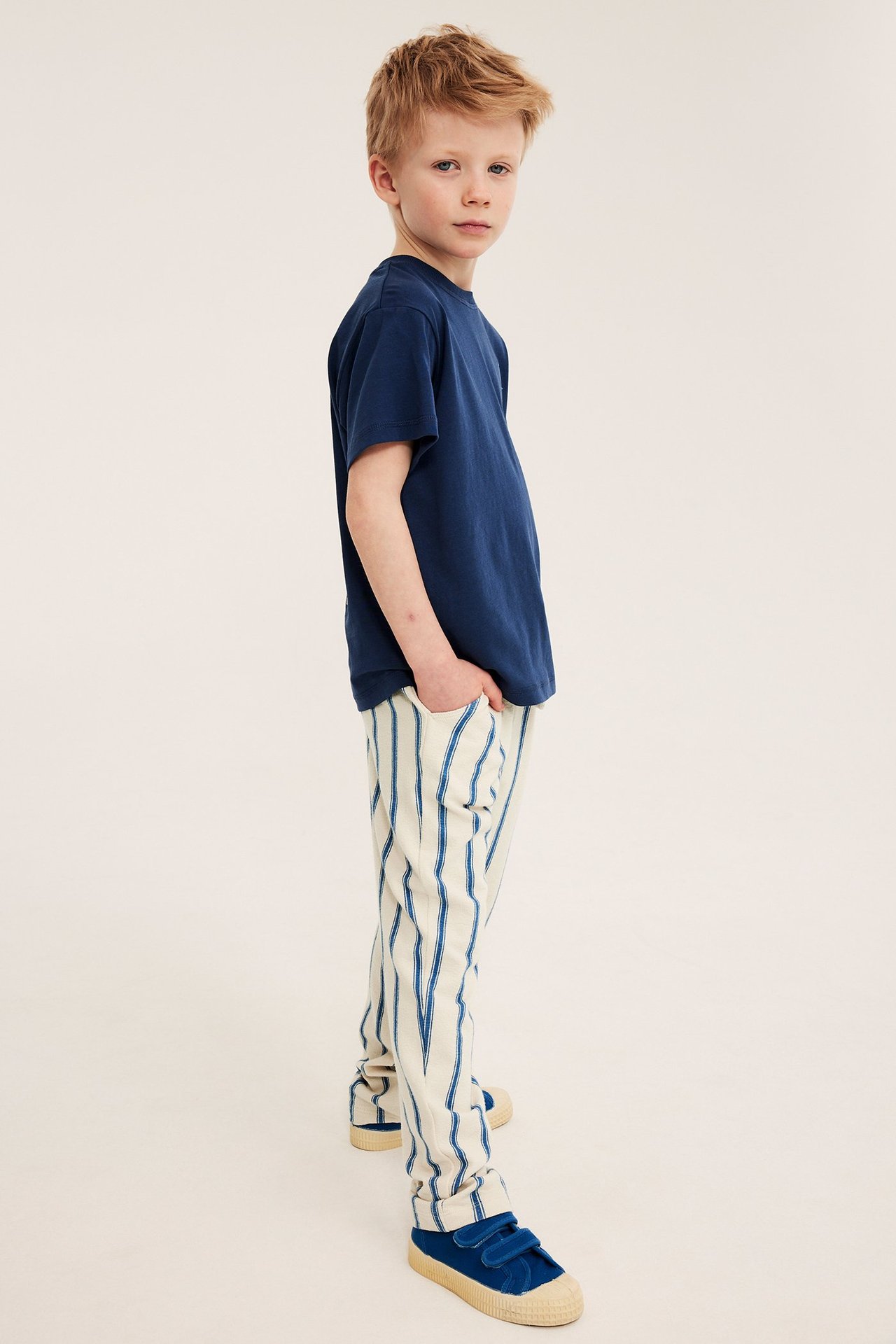 Widok z boku: Chłopiec w beżowo-niebieskich spodniach w paski, granatowej koszulce i niebieskich butach.