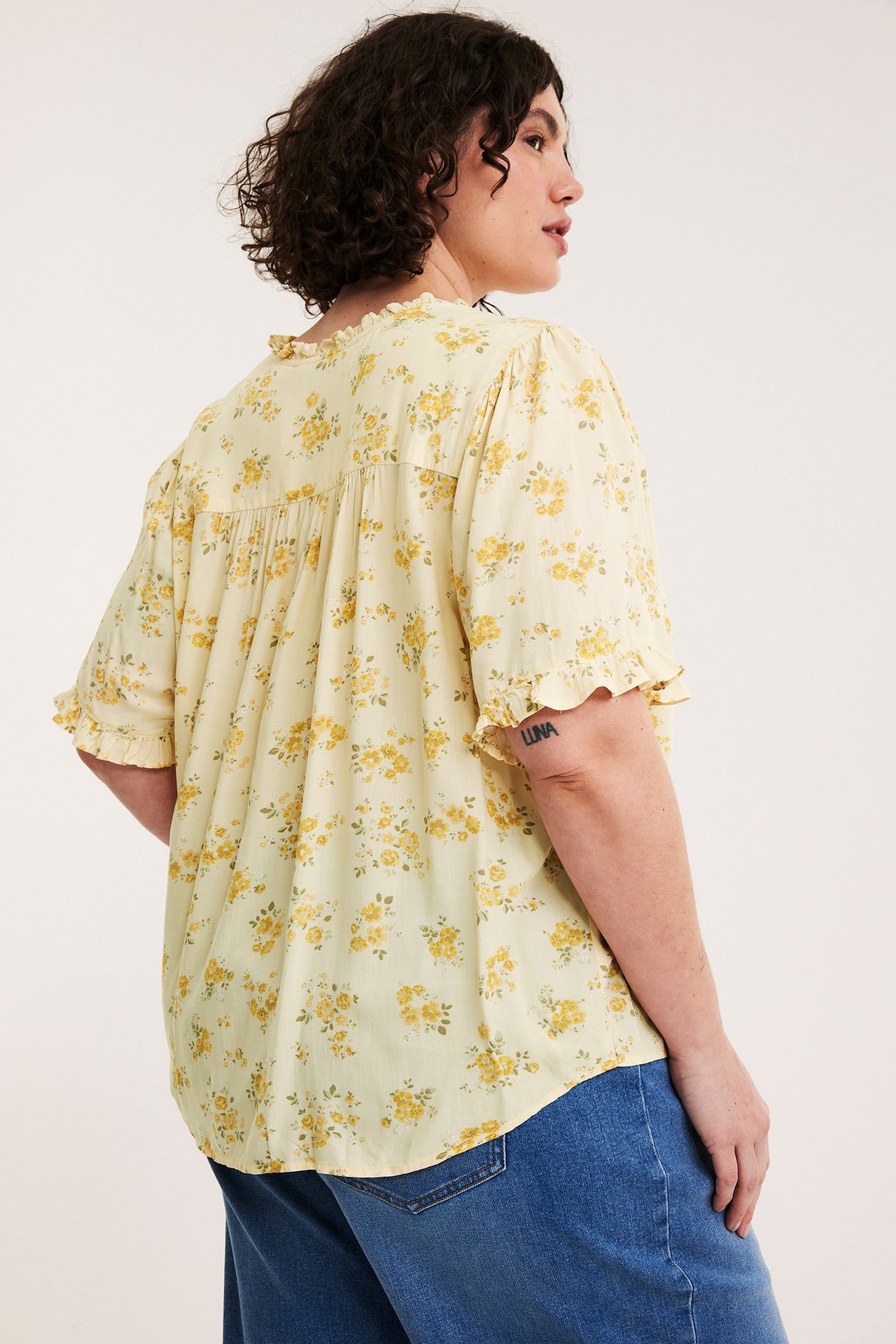 Kobieta tyłem: jasnożółta bluzka w kwiaty z falbanami przy dekolcie i rękawach, oraz niebieskie dżinsy.