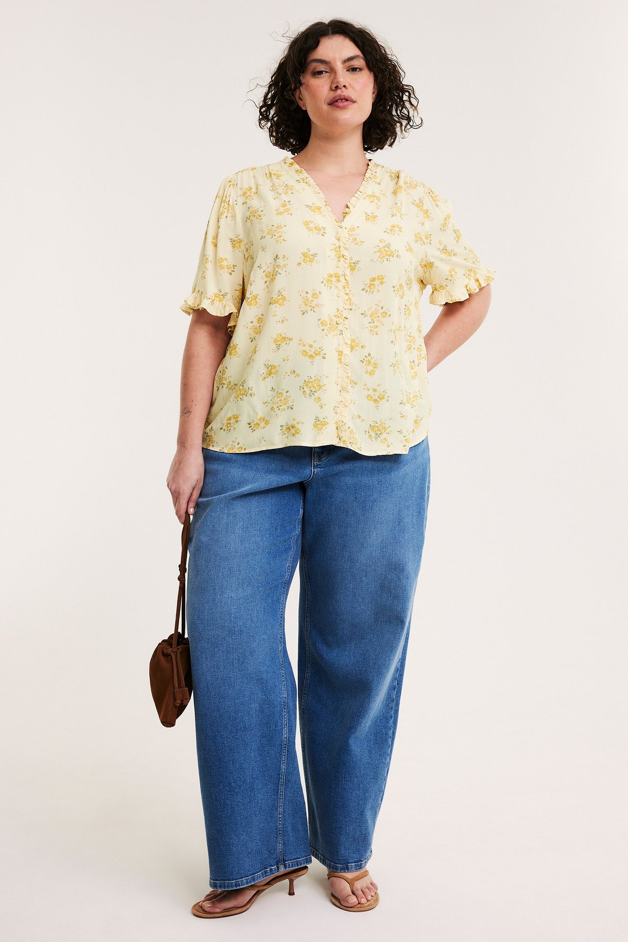 Frontvisning: Kvinne i lys gul blomstret volangbluse, blå vide jeans, brune sandaler, holder en brun veske.
