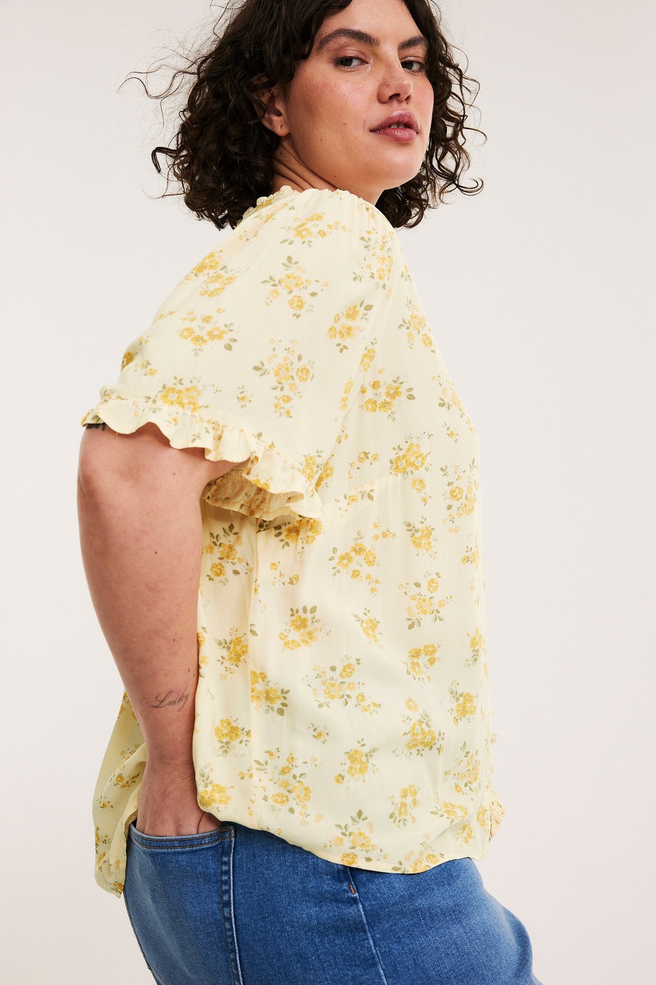 Widok z boku kobiety w jasnej żółtej bluzce w kwiaty z falbanami na rękawach i niebieskich jeansach.