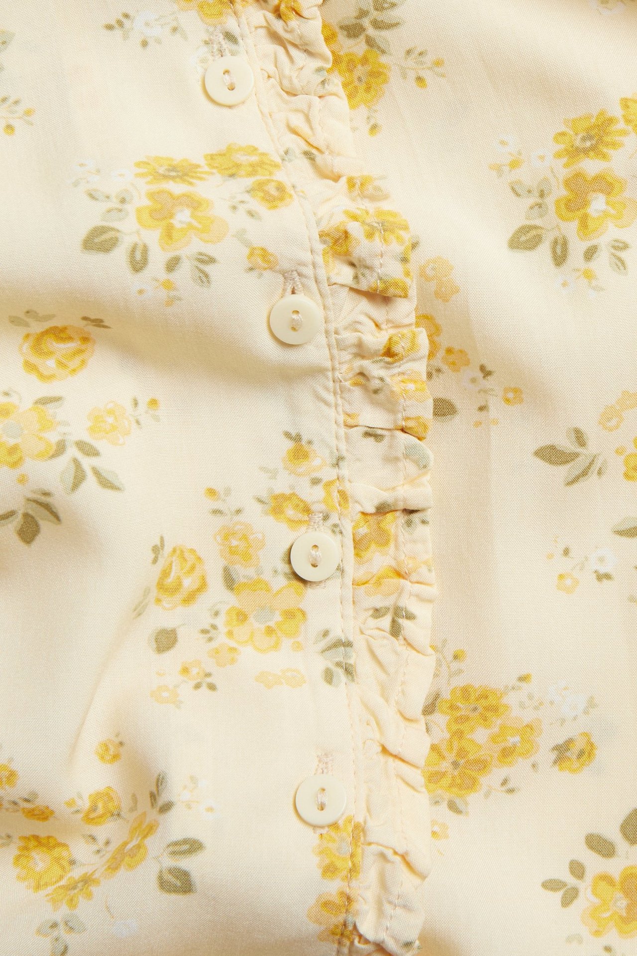 Zbliżenie na jasnożółtą wiskozową bluzkę w kwiatowy wzór, z krytymi guzikami i falbankami.