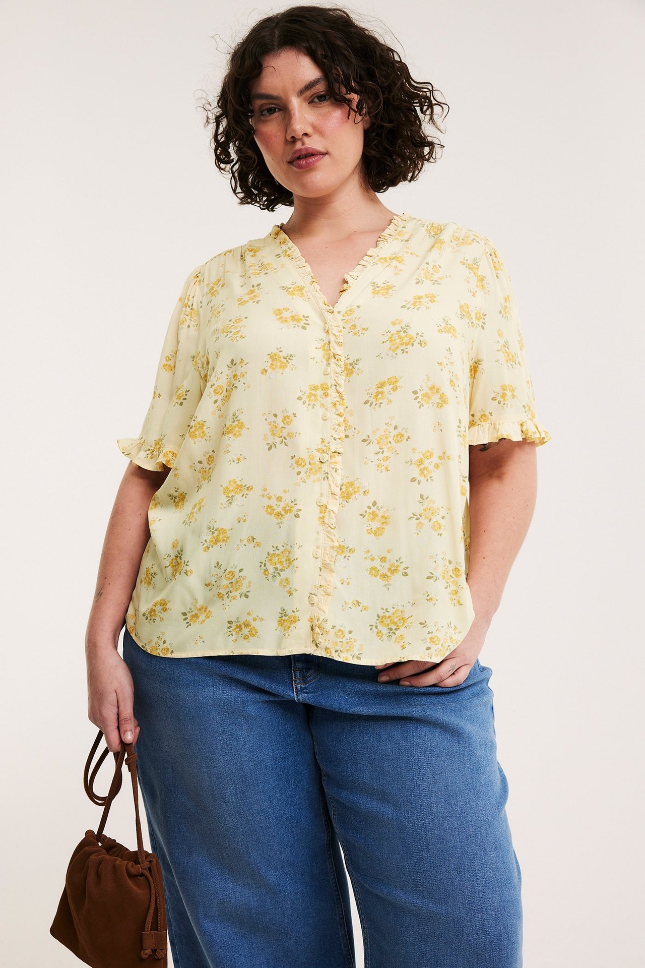 Widok z przodu: Kobieta w jasnożółtej bluzce w kwiaty z falbankami, niebieskich dżinsach, trzyma brązową torebkę.