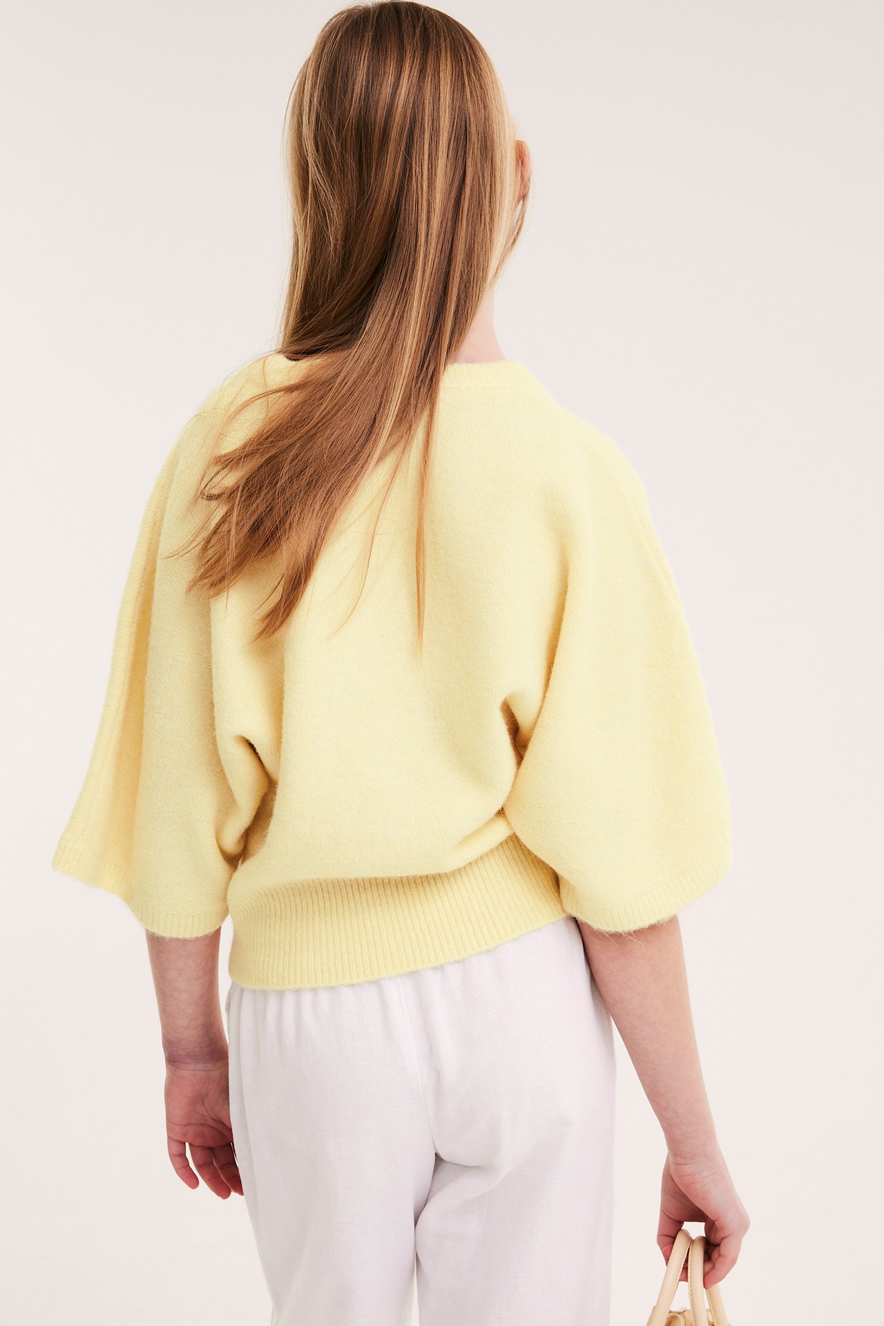 Widok z tyłu dziecka w jasnożółtym swetrze z szerokimi rękawami, białych spodniach, z małą plecioną torbą.