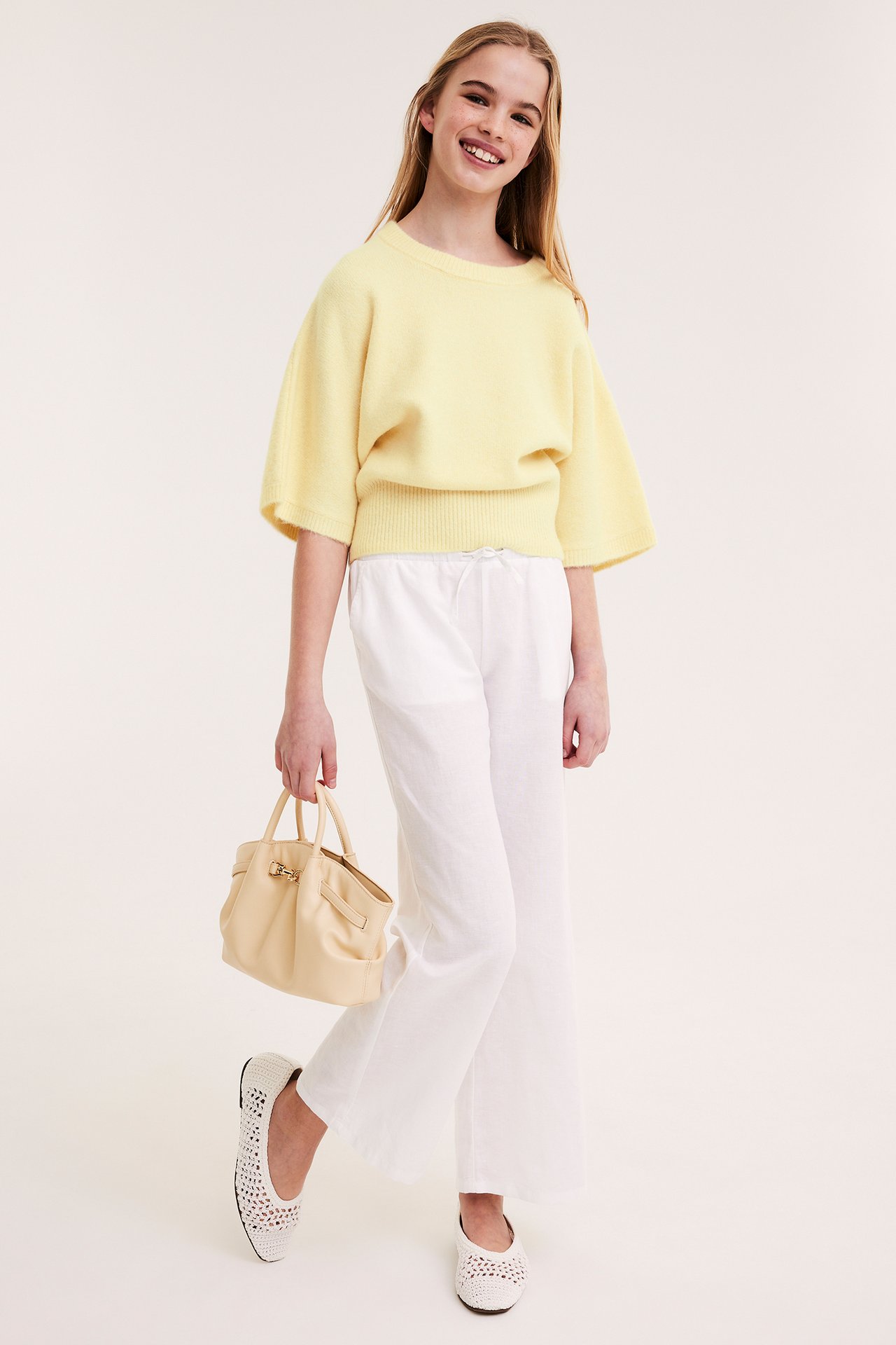 Smilende barn i lysegul strikket topp, hvite vide bukser, hvite ballerinasko, holder en lysegul håndveske.