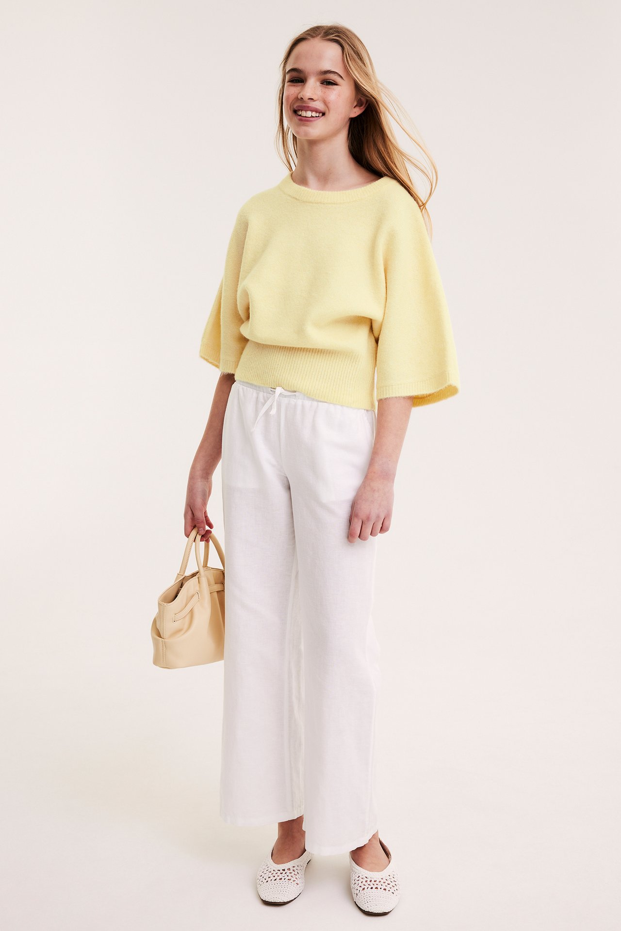 Dziecko ma na sobie jasnożółty dzianinowy top, białe szerokie spodnie, białe plecione baleriny, pasującą torebkę.