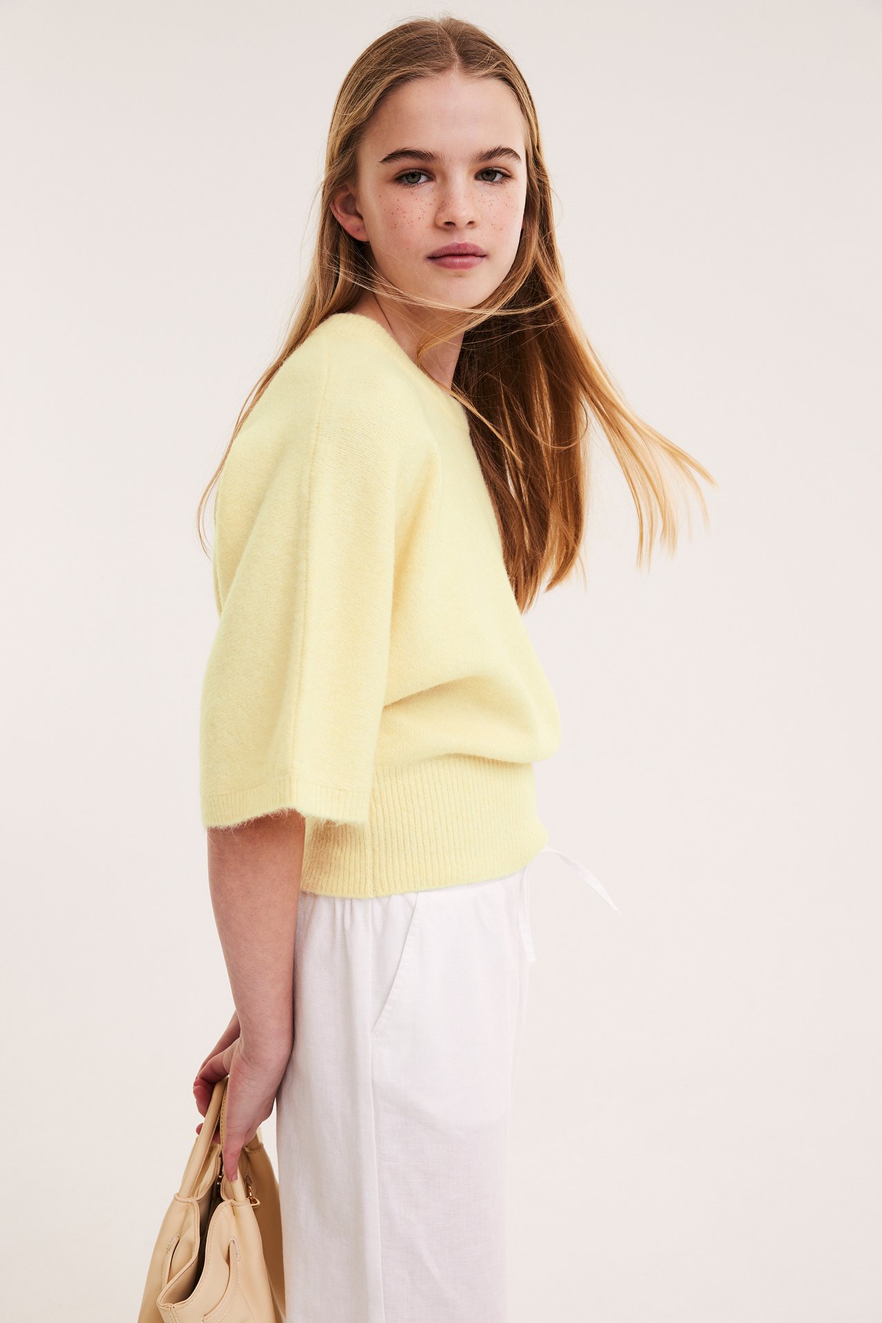 Widok z przodu-boku dziecka w jasnożółtym dzianinowym topie, białych spodniach, trzymającego beżową torebkę.