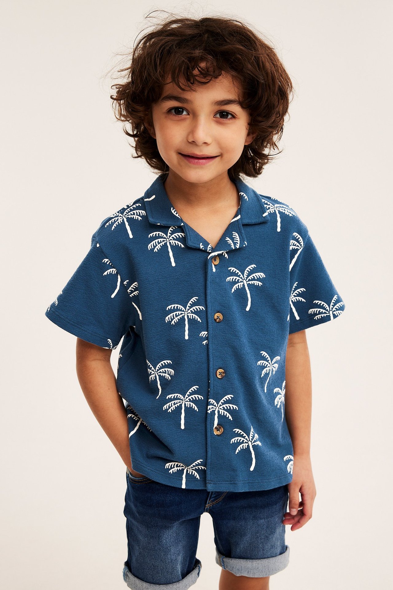 Frontvy av barn i blå relaxed fit skjorta med vitt palmträdsmönster och blåa denimshorts.