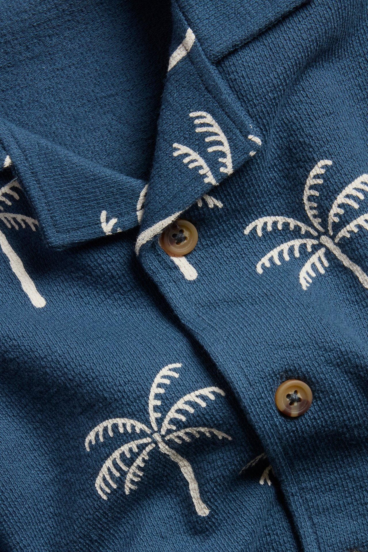 Niebieska bawełniana koszula z dżerseju, z białym wzorem palm i brązowymi guzikami.