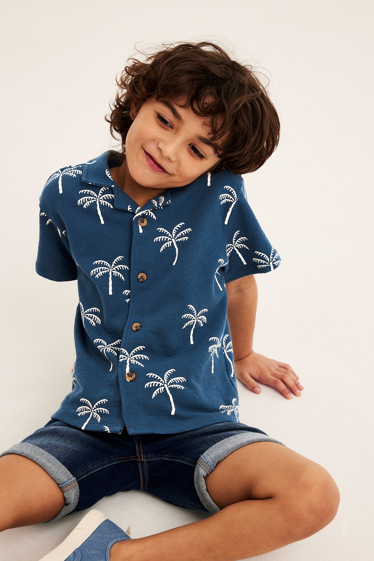 Frontvy av ett barn i blå kortärmad bomullsskjorta med vitt palmprint och mörkblå jeansshorts.