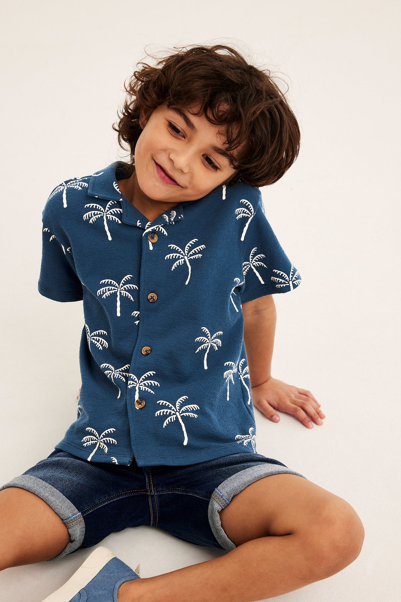 Frontvy av ett barn i blå kortärmad bomullsskjorta med vitt palmprint och mörkblå jeansshorts.