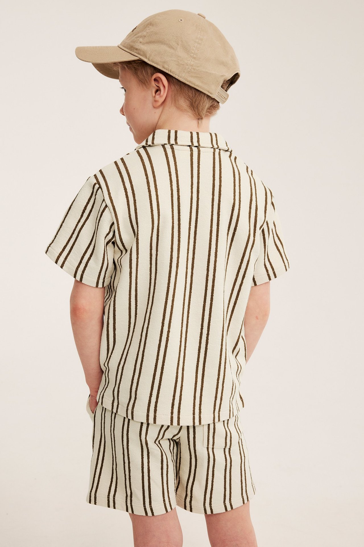 Bakifrån på ett barn i en ljusbeige randig bomullstrikåskjorta, matchande shorts och keps.