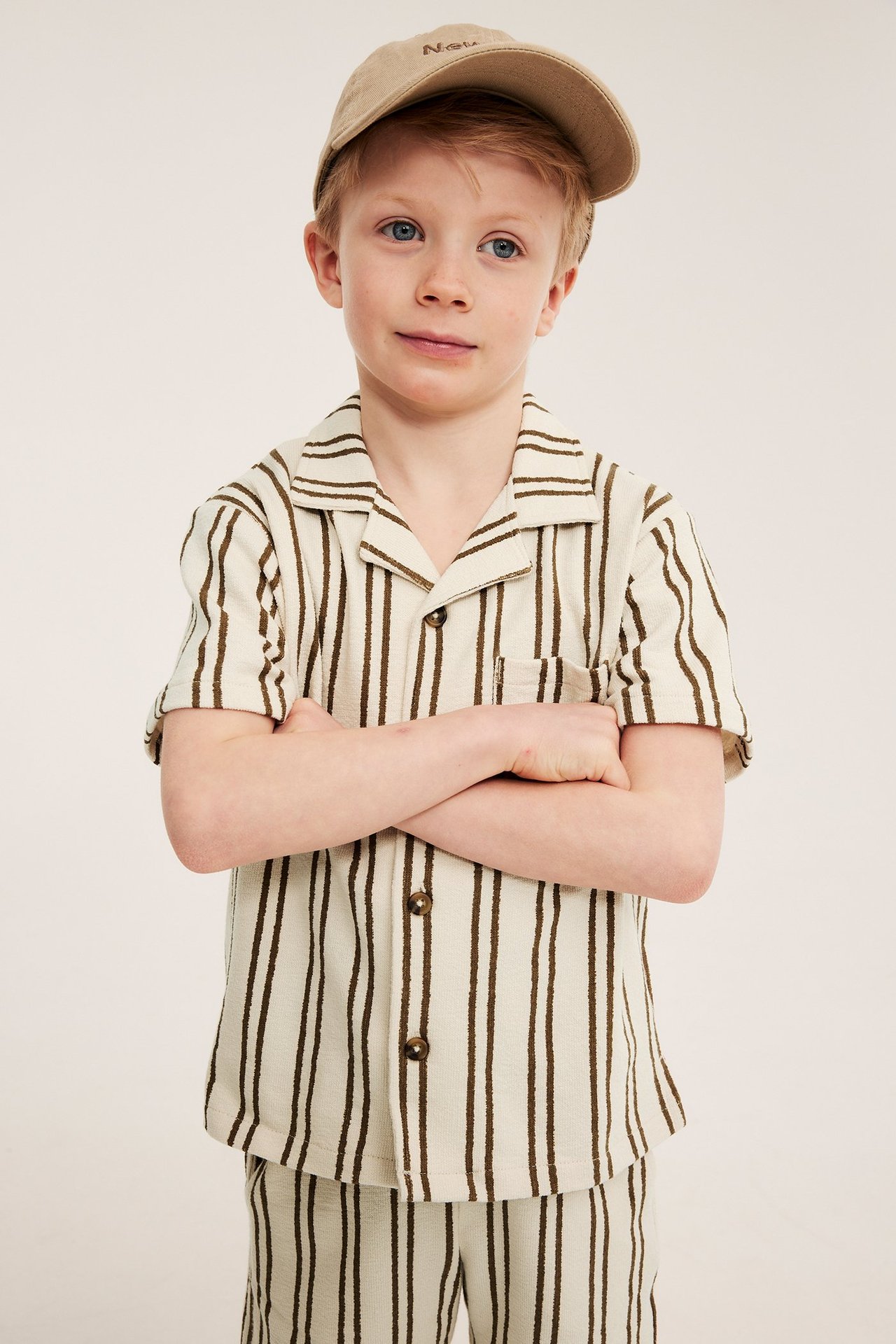 Front view: Barn bär ljusbeige randig bomullstrikåskjorta med knappar, matchande shorts och beige keps.