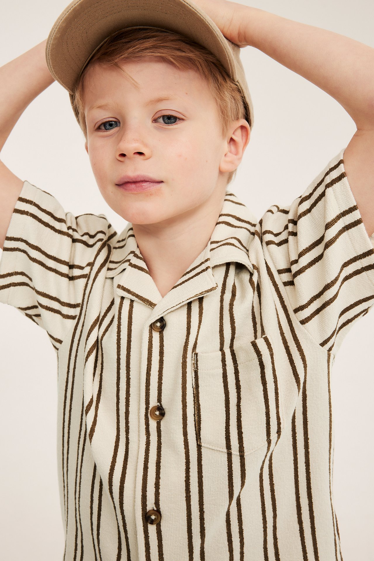 Barn bär ljusbeige randig kortärmad bomullsskjorta med bröstficka och beige keps.