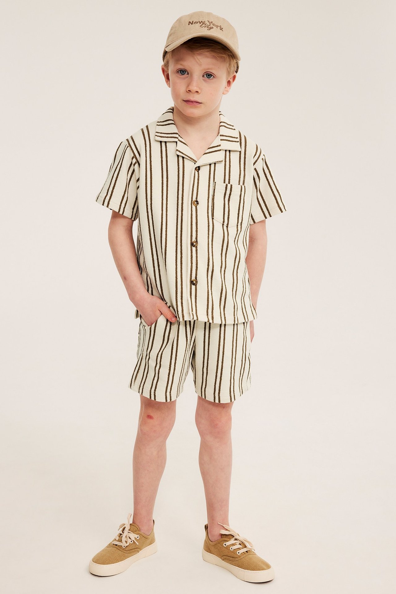 Pojke bär en ljusbeige och brunrandig skjorta i bomullstrikå, matchande shorts, en beige keps och sneakers.