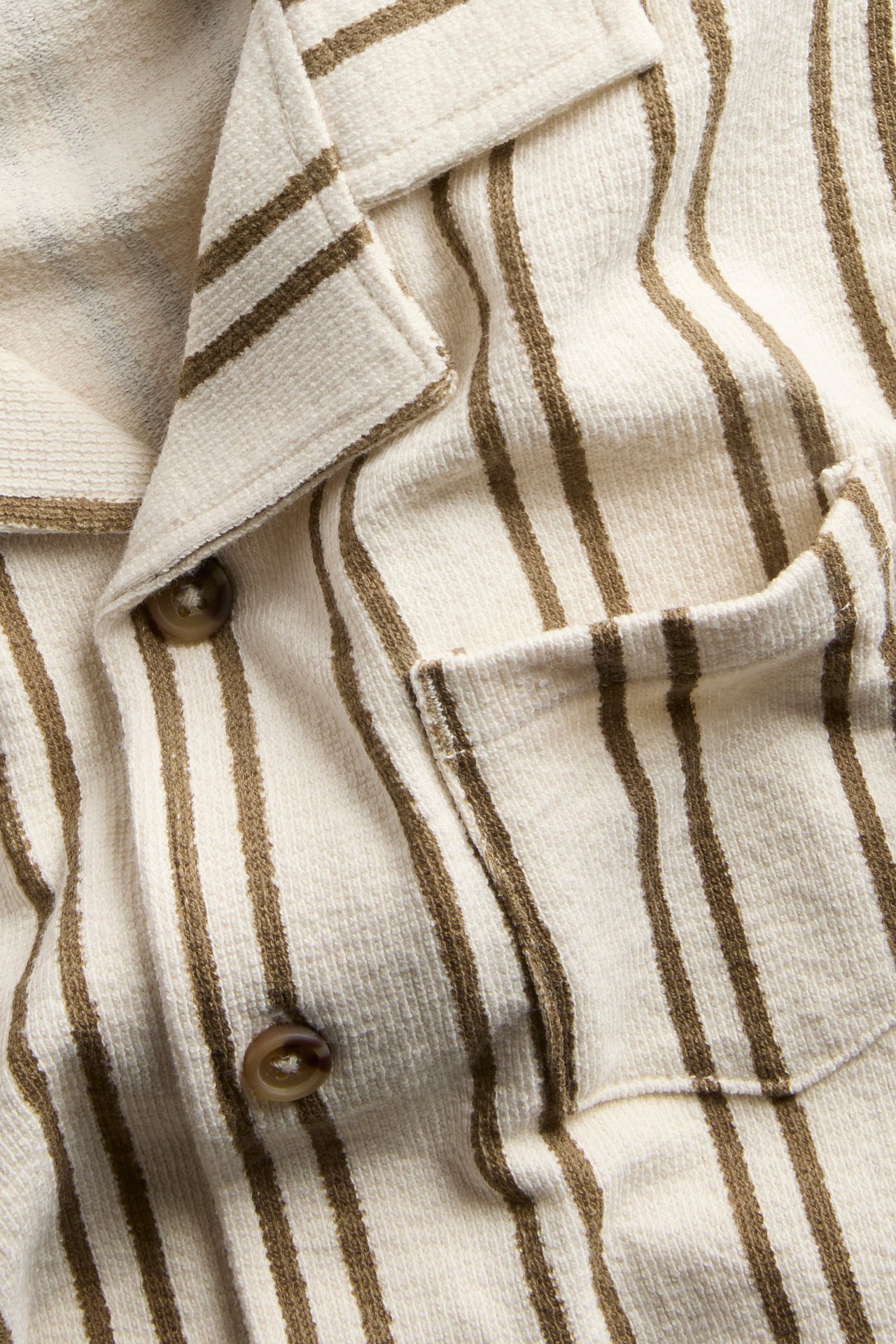 Närbild framifrån på en ljusbeige bomullstrikåskjorta med bruna ränder, bröstficka och bruna knappar.