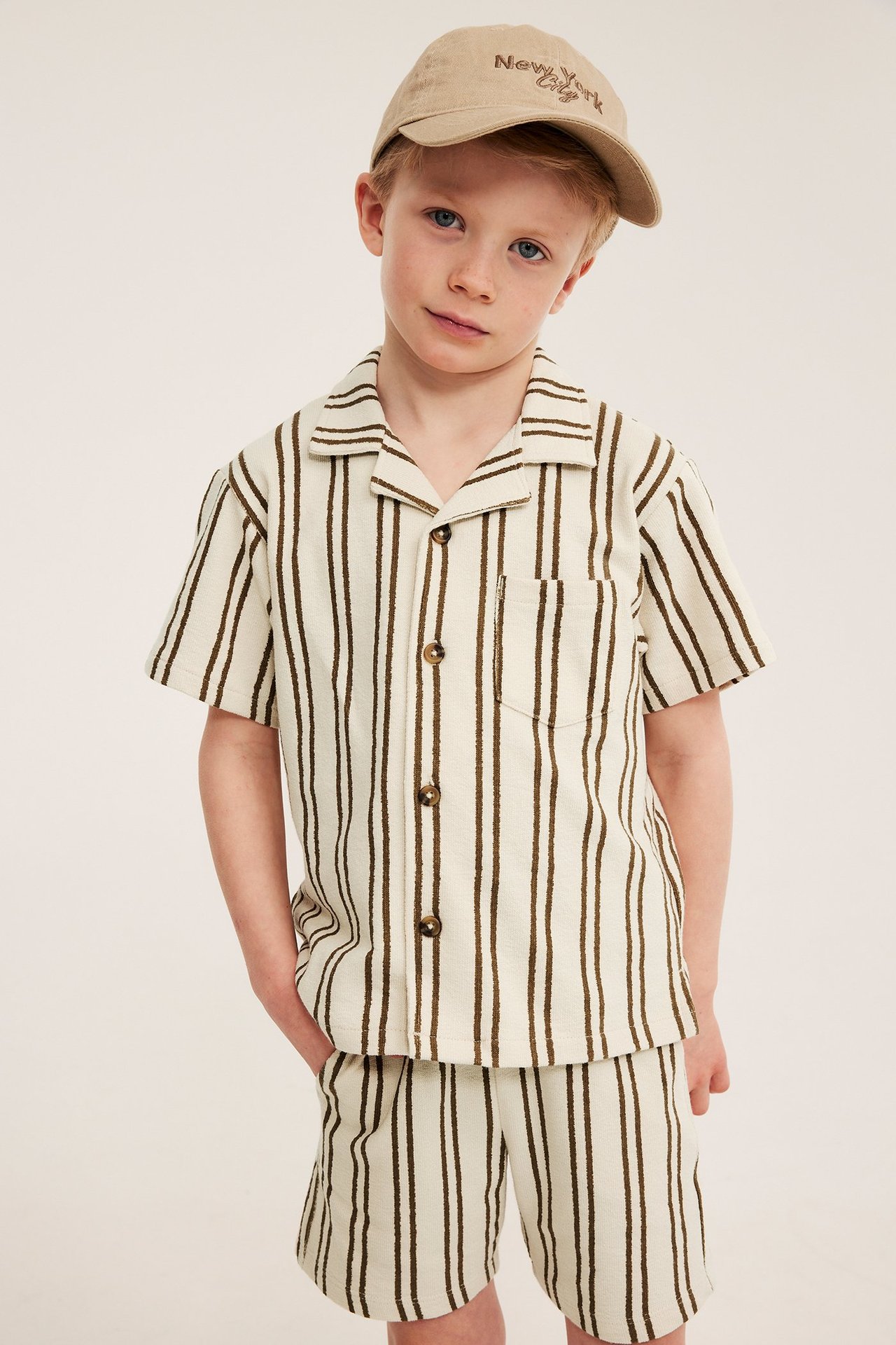 Framifrån: Ett barn i ljusbeige randig bomullstrikåskjorta, matchande shorts och beige keps.