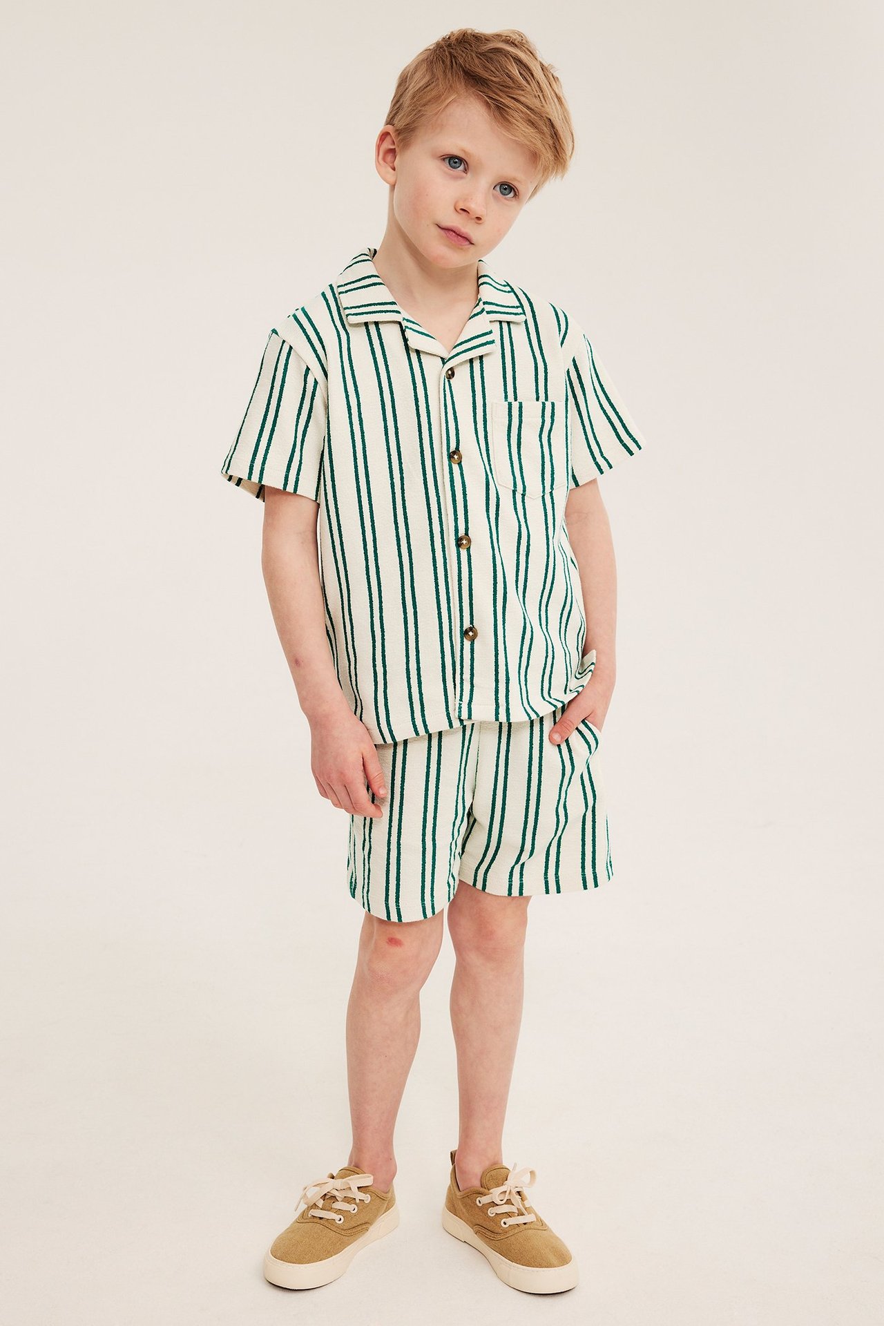 Frontvy av ett barn i lös gräddvit och grönrandig kortärmad skjorta, matchande shorts och beiga sneakers.