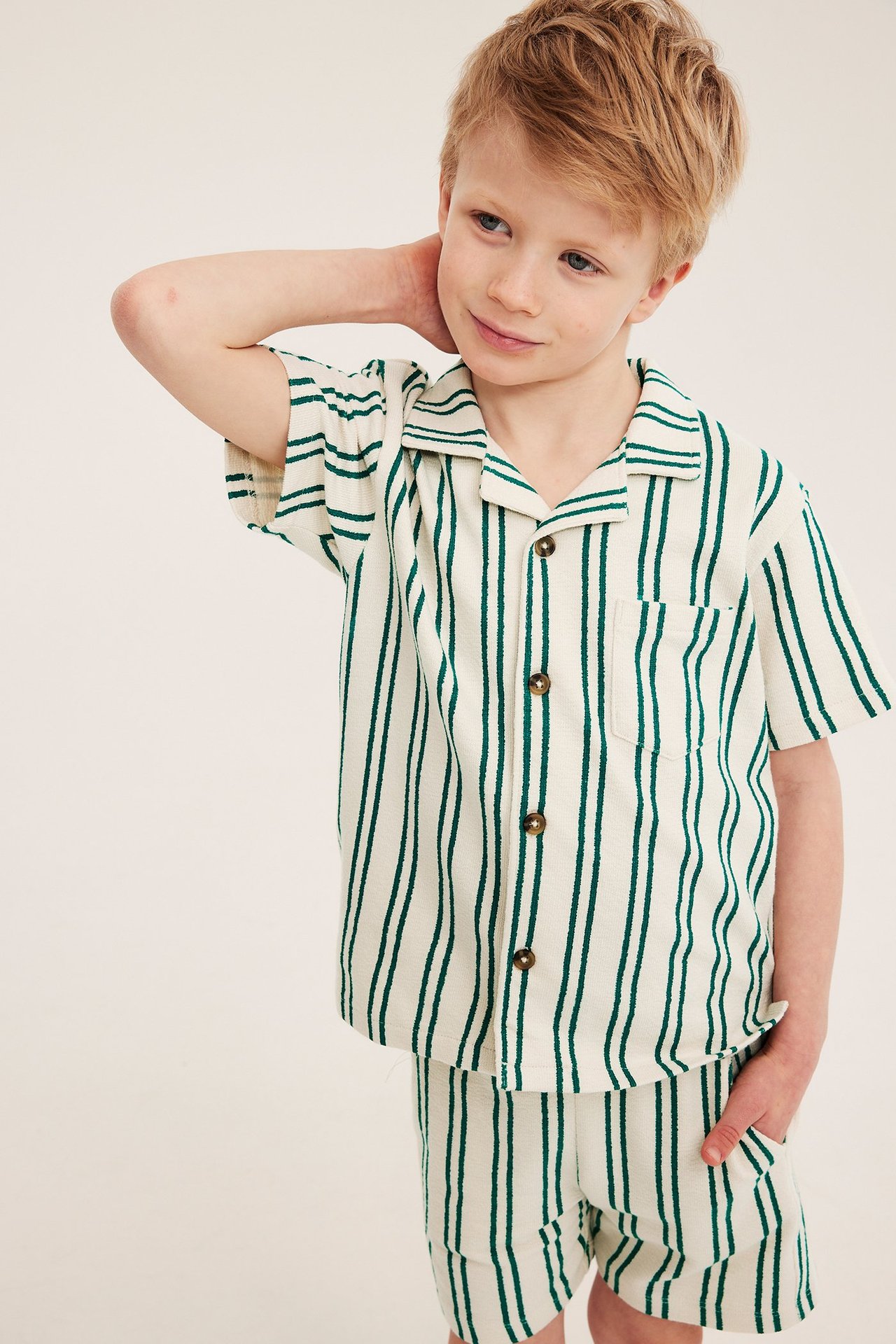 Framifrån: Ett barn bär en lös grön- och cremefärgad randig bomullsskjorta och matchande shorts.