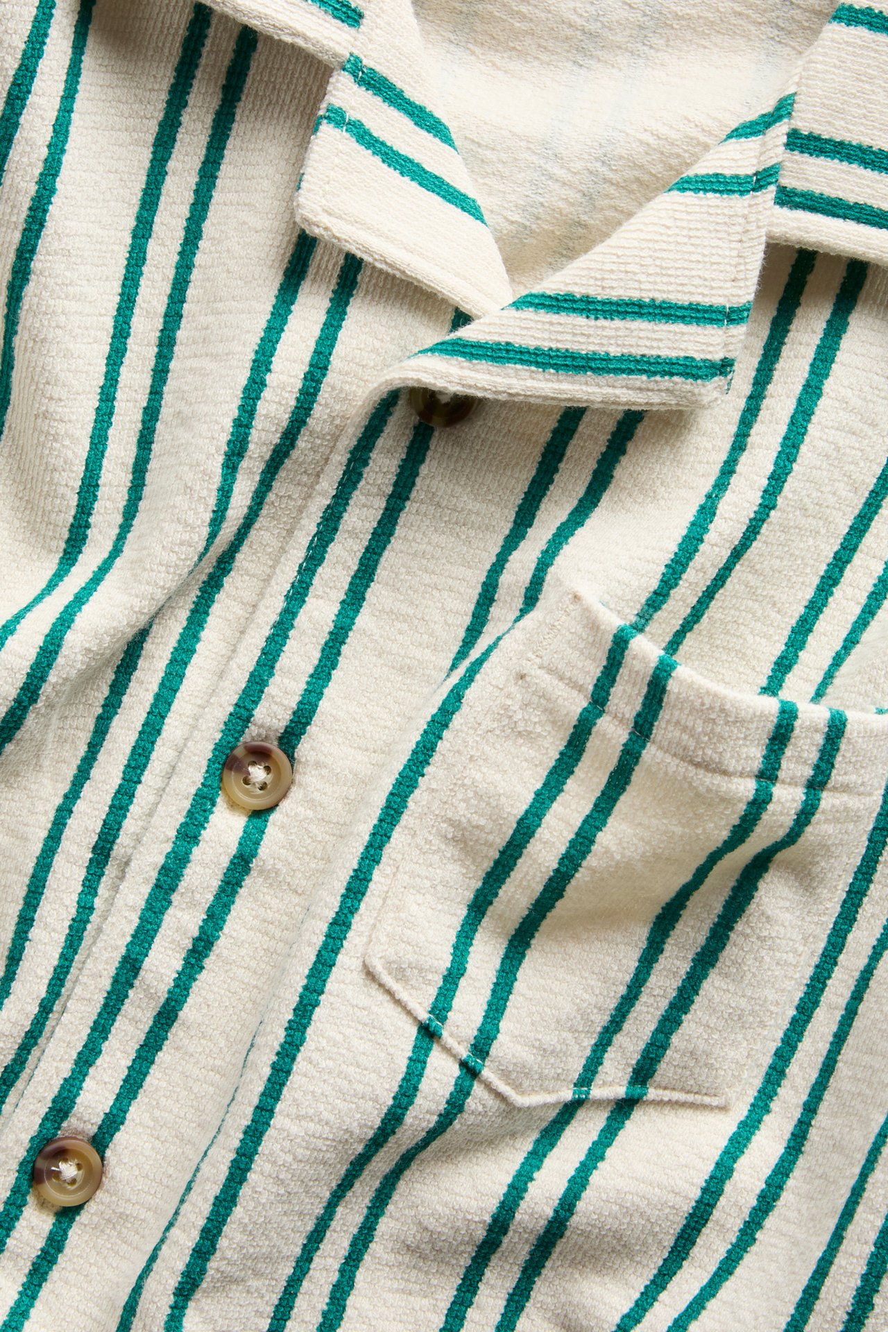 Närbild, framifrån: en offwhite bomullstrikåskjorta med gröna ränder, bruna knappar och bröstficka.