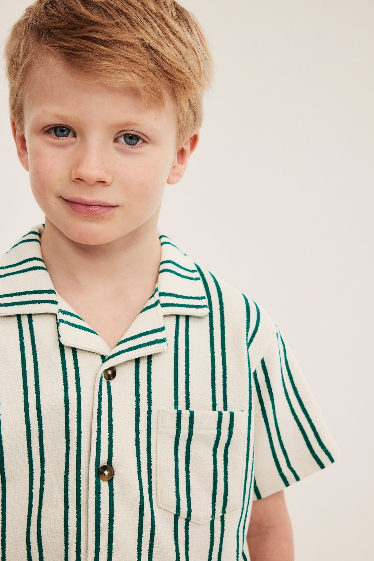 Närbild framifrån: Kid i gräddvit trikåskjorta med gröna ränder, knappar fram och bröstficka.
