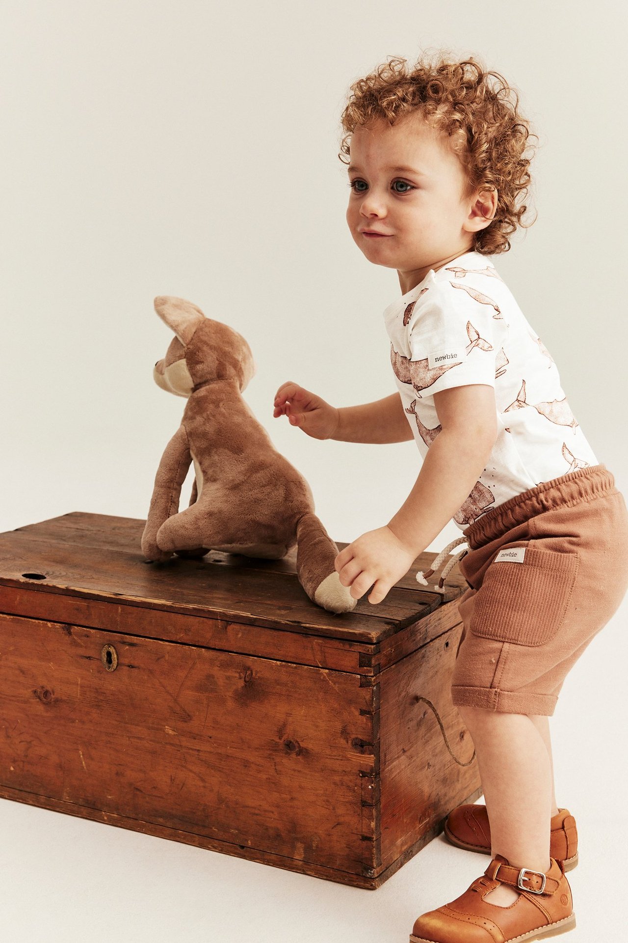 Fra siden, baby i en offwhite kortarmet body med hvalmønster, brune shorts og brune spennesko.