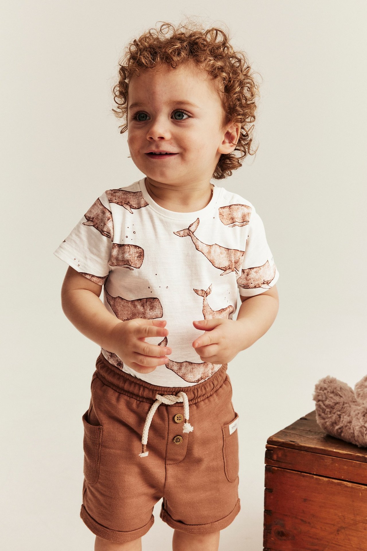 Widok z przodu: niemowlę w ecru bawełnianym body z waleniami i brązowych szortach.