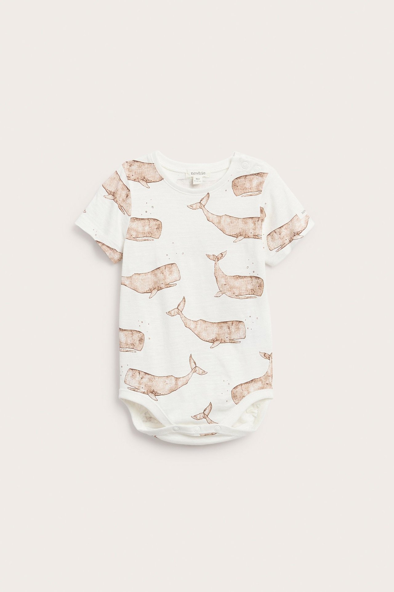 Przód białego bawełnianego body niemowlęcego z krótkim rękawem, z brązowym wzorem w wieloryby i zatrzaskami.