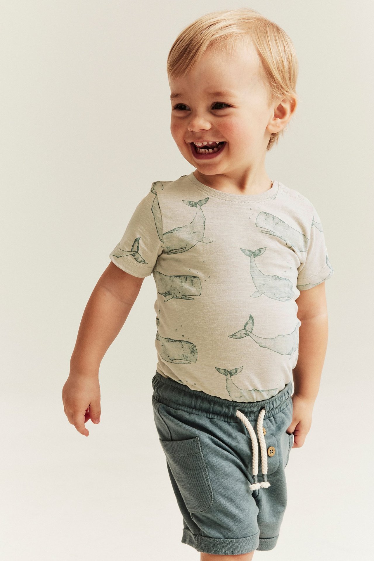 Widok z przodu uśmiechniętego dziecka w beżowym body z krótkim rękawem w wieloryby i ciemnozielonych szortach.