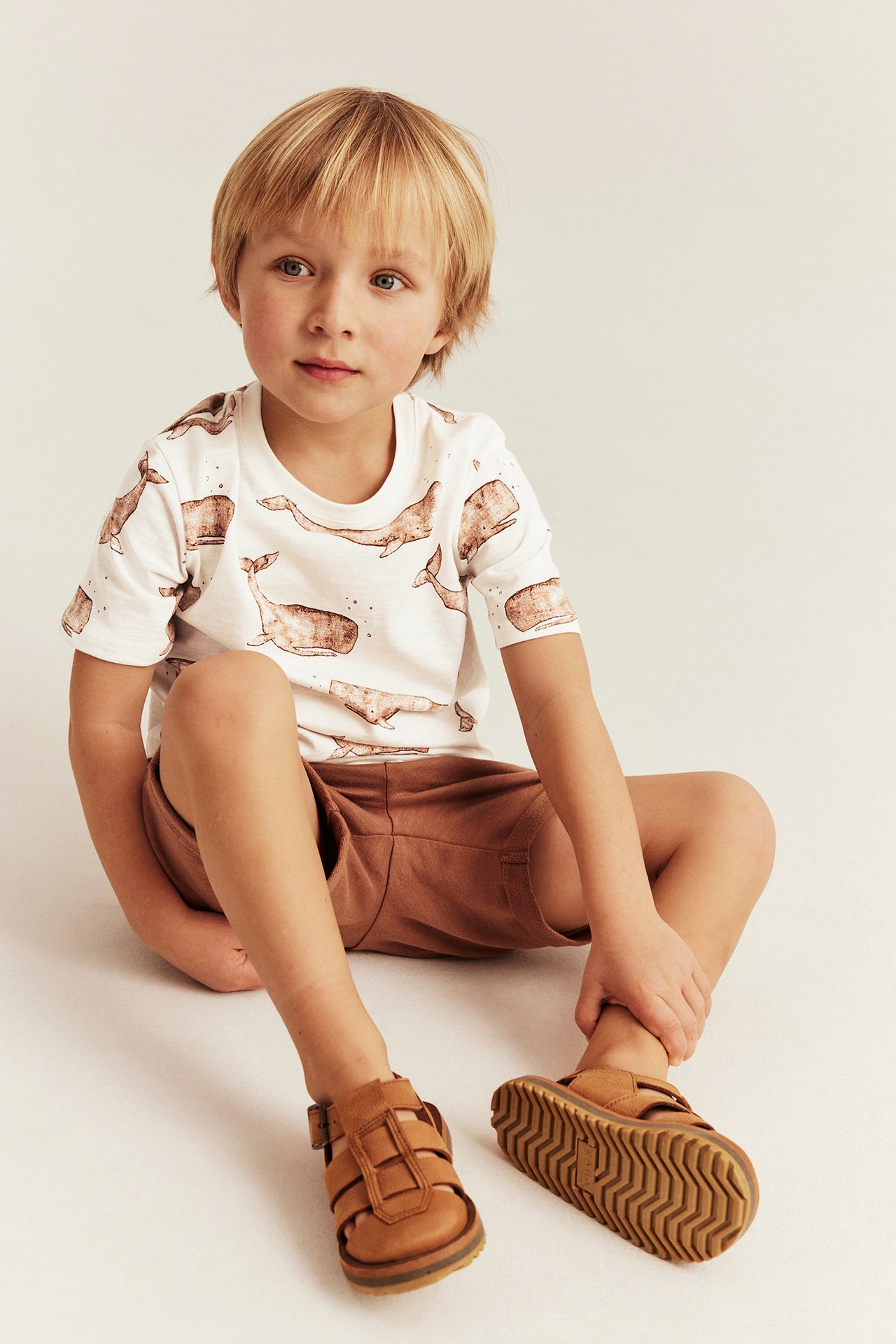 Widok z przodu: dziecko w białym bawełnianym t-shircie ze wzorem w wieloryby, brązowych szortach i brązowych sandałach.