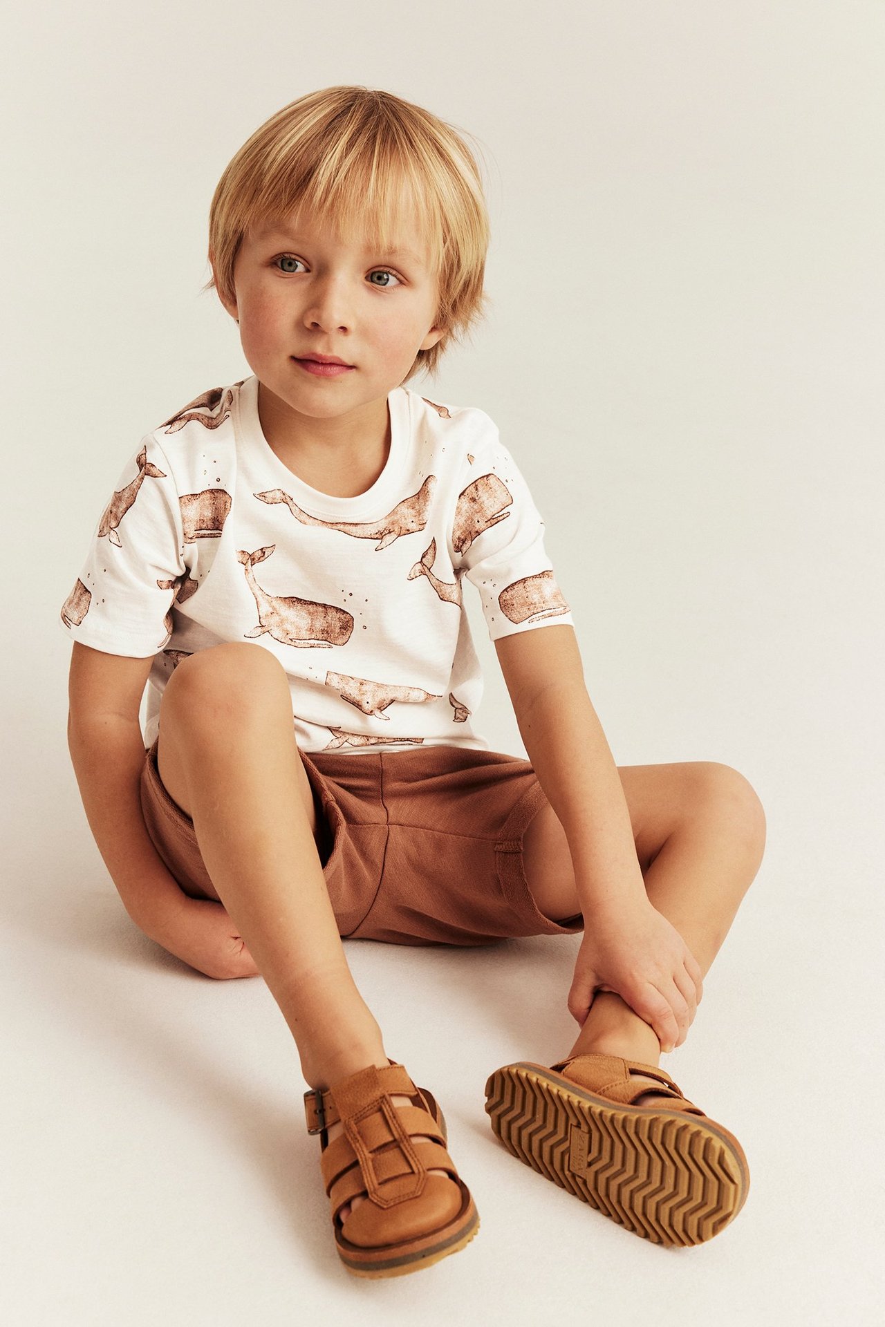 Widok z przodu: dziecko w białym bawełnianym t-shircie ze wzorem w wieloryby, brązowych szortach i brązowych sandałach.