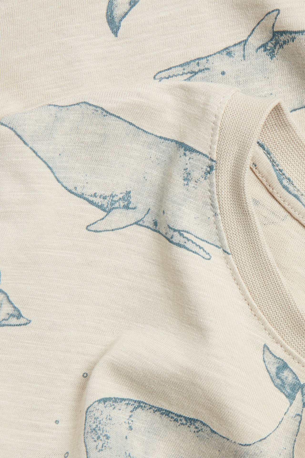 Nærbilde av en beige slub-jersey t-skjorte med blått hvalmønster og ribbet halskant.