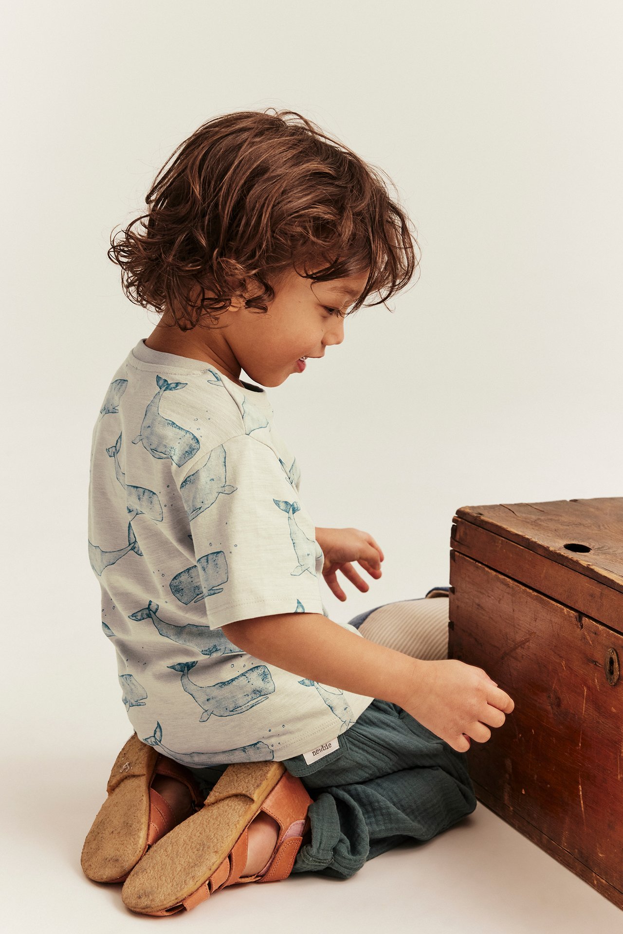 Widok z boku dziecka w beżowej koszulce slub z wzorem wielorybów, ciemnozielonych spodniach i brązowych sandałach.